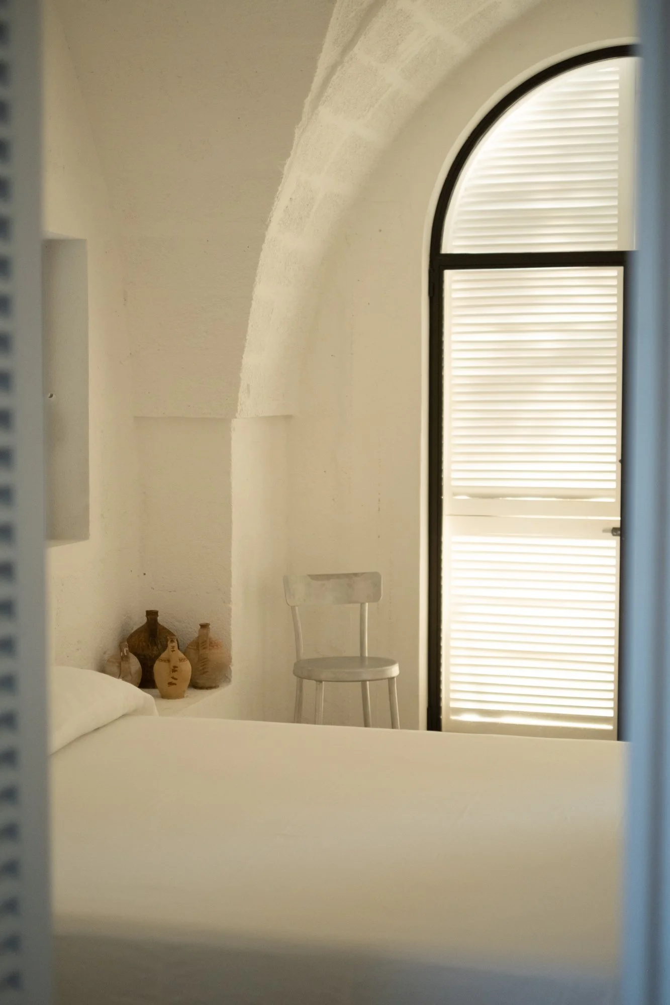 A cozy bedroom with a white bed, decorative vases on a small shelf, a white chair, and a large arched window with closed blinds.