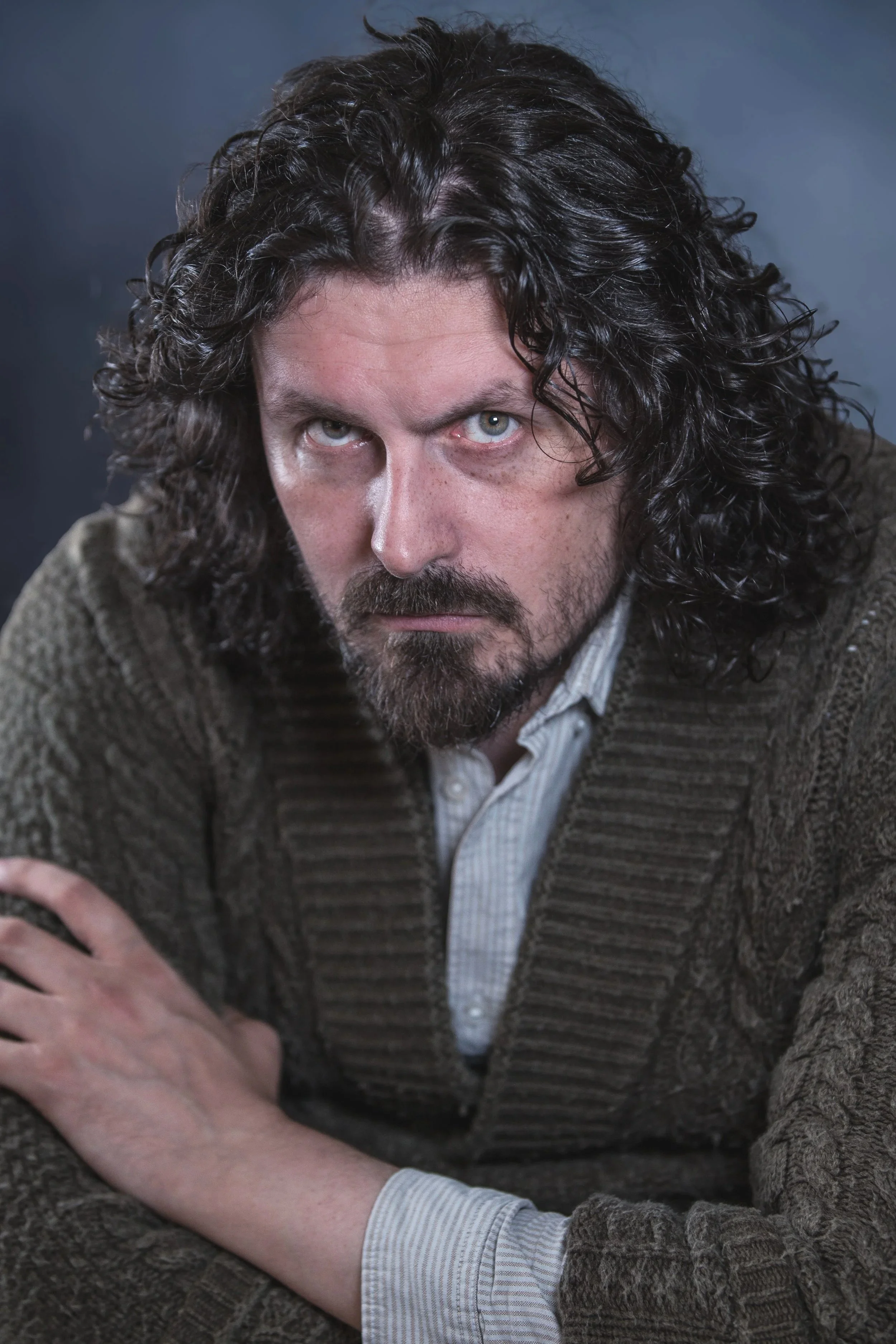 Close-up portrait of a man with long, curly dark hair and a beard, wearing a brown textured sweater over a light-colored shirt, looking intently at the camera against a dark background.