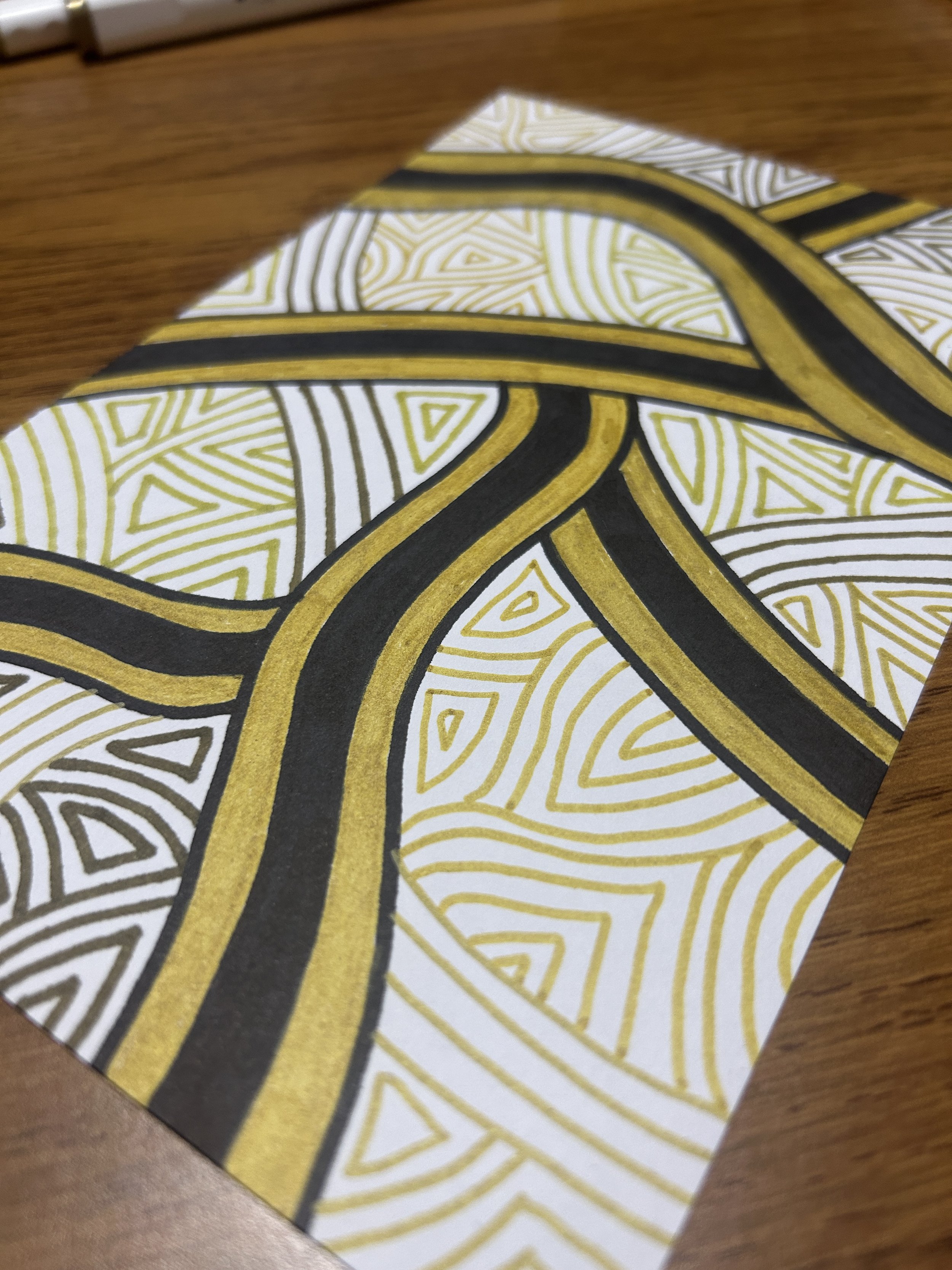 Close-up of a colorful, patterned paper with black, gold, and white lines forming intricate geometric designs, placed on a wooden surface.
