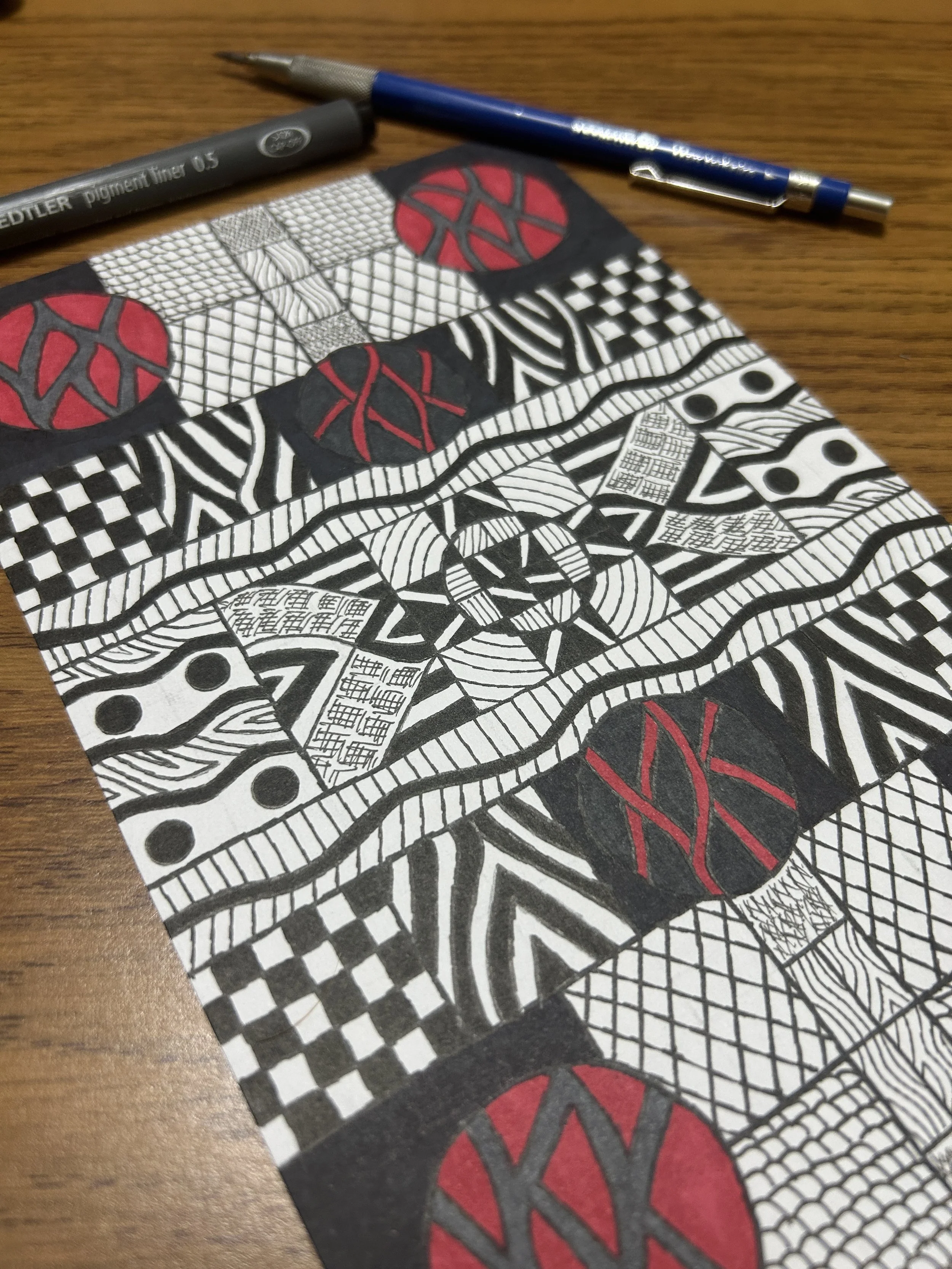 Black and white abstract geometric drawing on paper, with red and black circles and patterns, resting on a wooden surface, alongside a grey pen and a blue mechanical pencil.