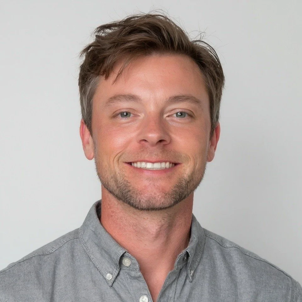 Portrait of a smiling Brandon Garrett - Founder of Refuge Roofing with brown hair, blue eyes, and light stubble, wearing a gray collared shirt, against a plain light background.