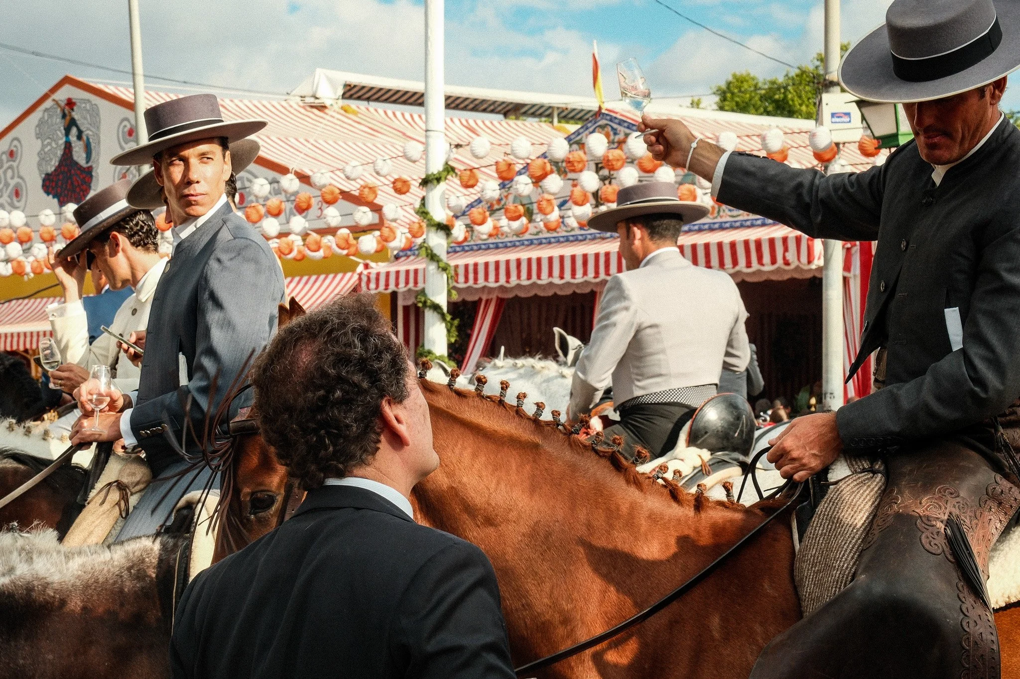 Groupe d'hommes en costumes traditionnels andalous, certains à cheval, lors d'une fête avec stands décorés de ballons et rubans rouges et blancs.