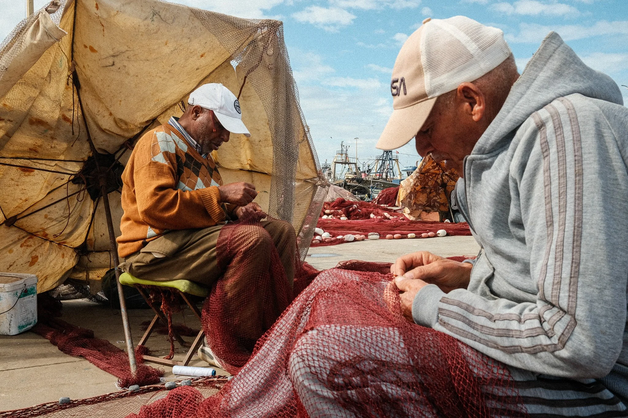 Deux hommes pêcheurs réparent des filets de pêche rouge en bord de mer, avec des bateaux de pêche en arrière-plan.