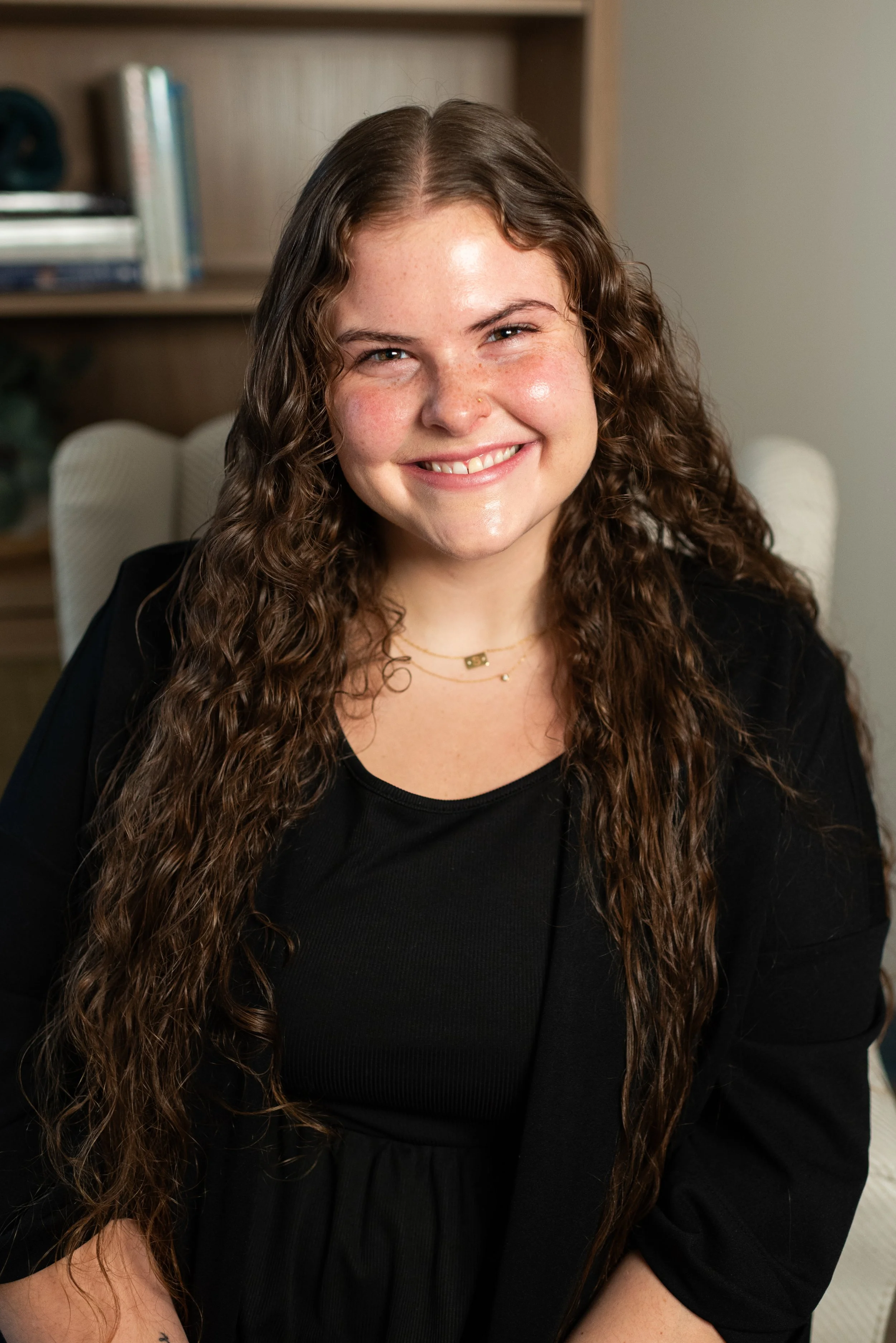 Young woman with long, curly hair smiling in front of a plain wall.