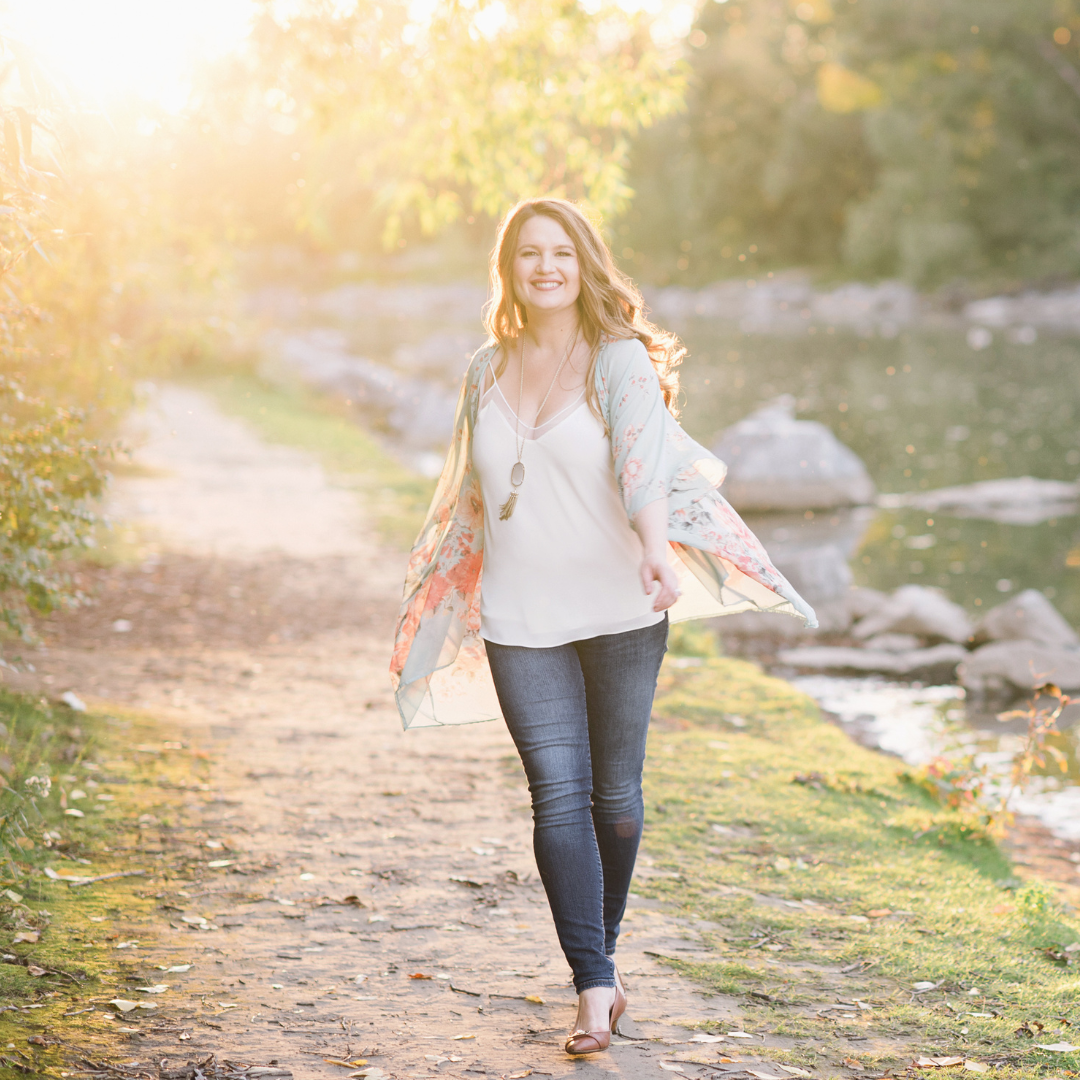 A woman smiling and walking along a dirt path by a river at sunset, wearing jeans, a white top, a floral kimono, and heels.