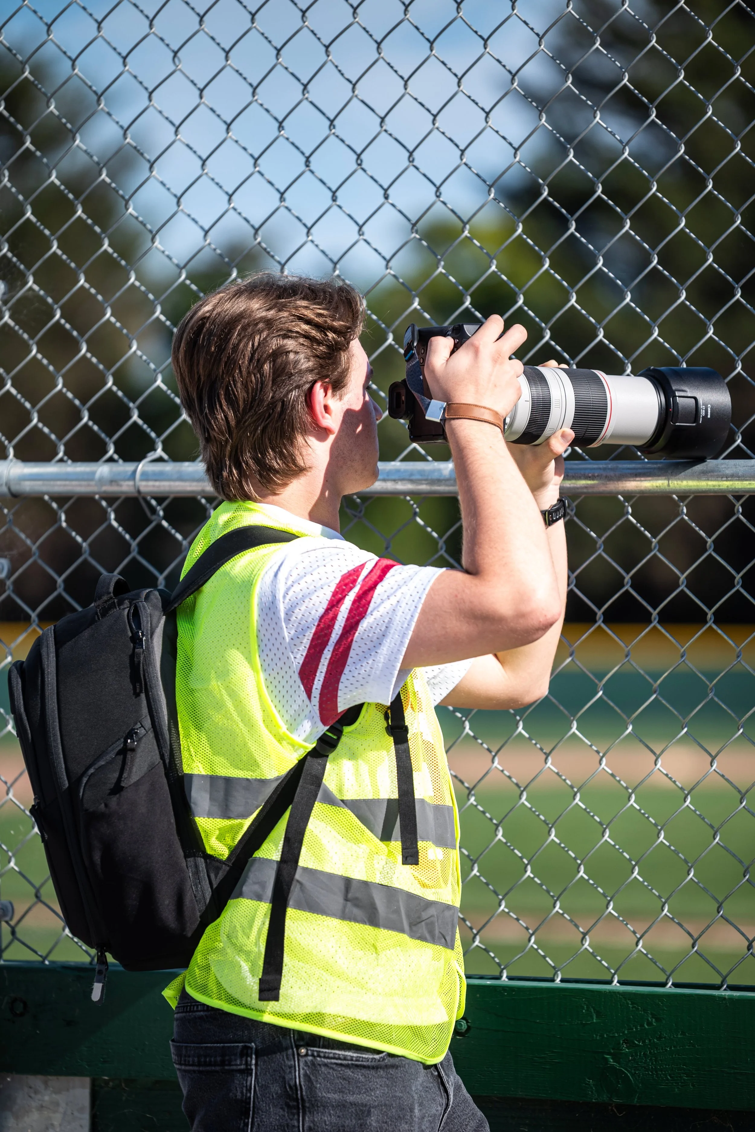 A person with brown hair wearing a yellow safety vest and black backpack takes a photo with a camera featuring a large telephoto lens, standing in front of a chain-link fence with a park or field in the background.