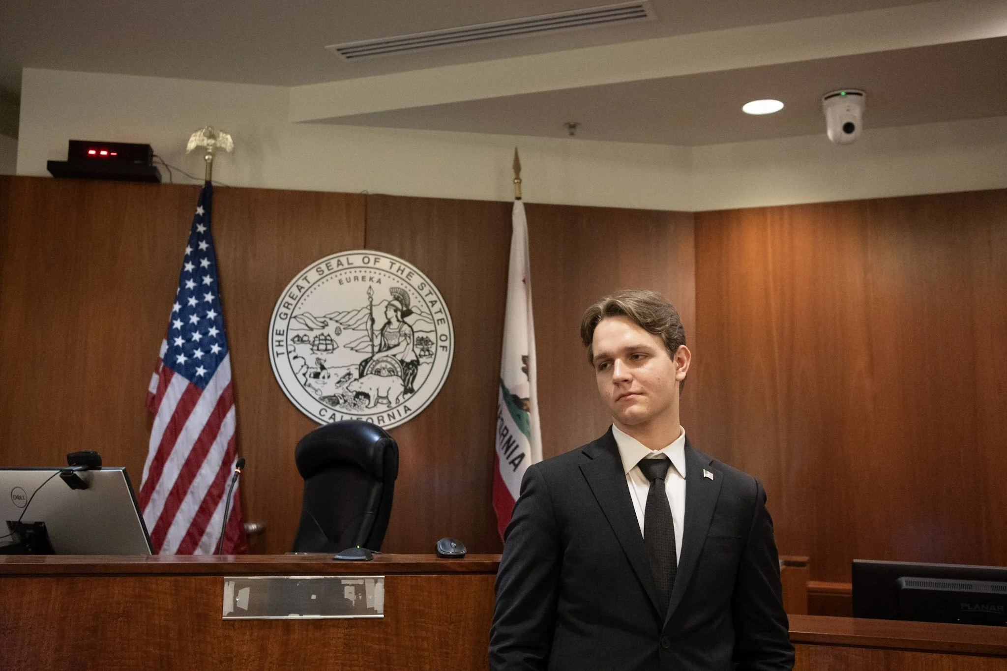 A young man in a black suit standing in a courtroom or government meeting room, with the California state seal and the American flag behind him.