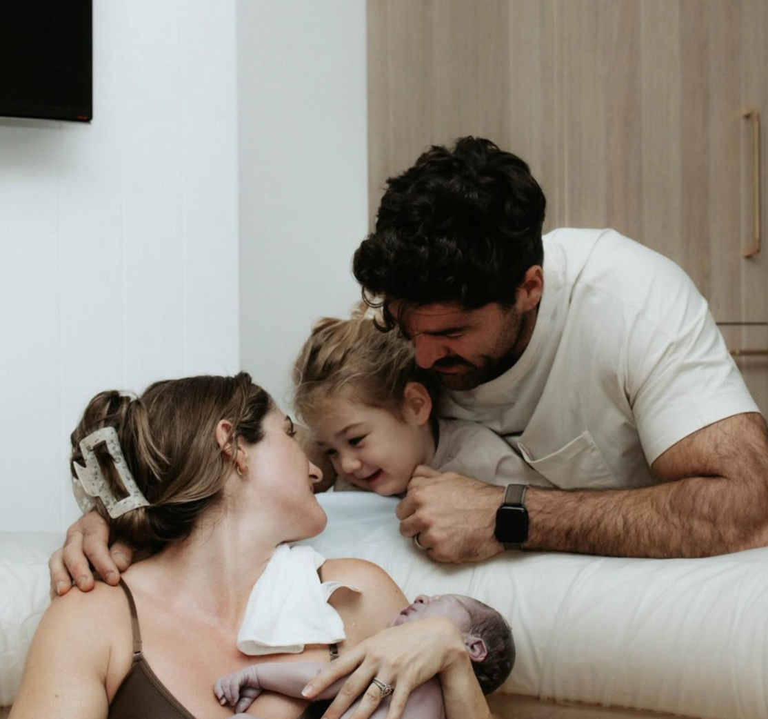 A woman who took HypnoBirthing classes Whittier, CA with Nurture The Divine holds her newborn and smiles at her family.