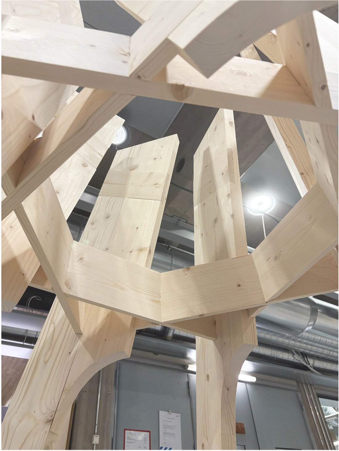 Interior view looking upward at unfinished wooden structure, possibly a chair or table frame, with visible wood grain and joints, in an industrial or workshop setting with exposed ducts and ceiling lights.