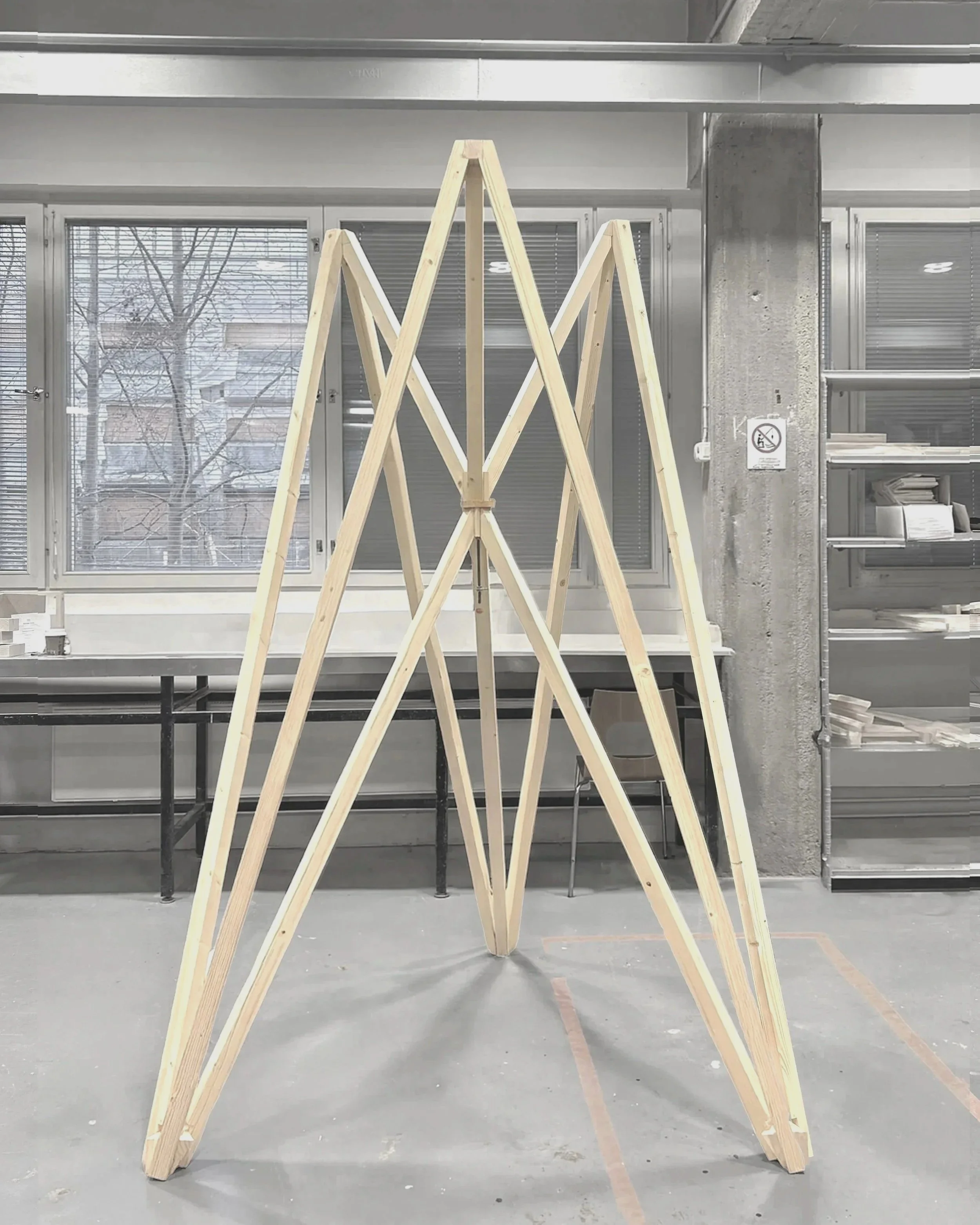 Interior view of a workshop with wooden tripod structures, large windows with blinds, shelves with papers, and a concrete pillar with a no smoking sign.