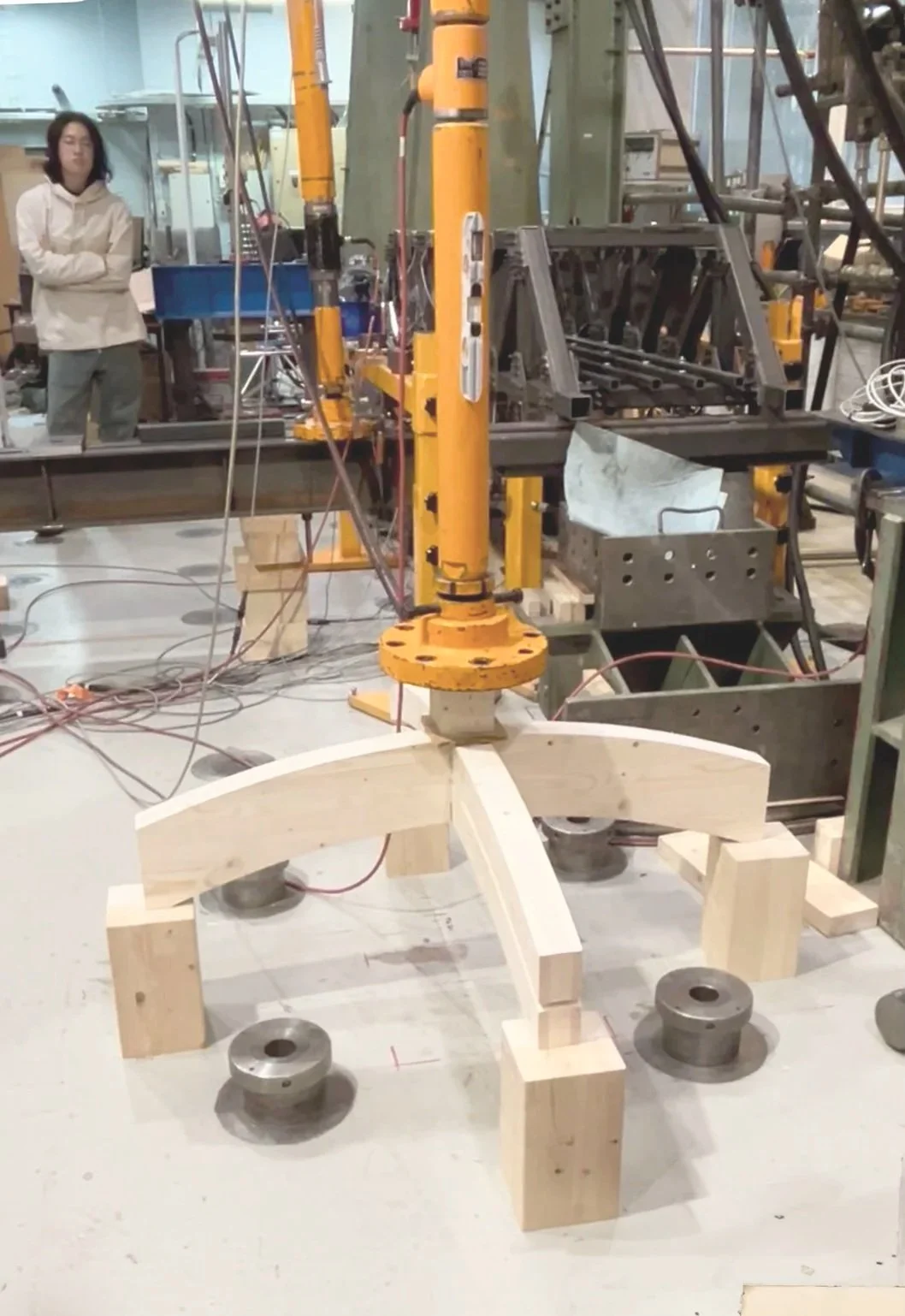 A person standing in a workshop with machinery, wooden components, and metal parts, working on a robotic arm or machine assembly.