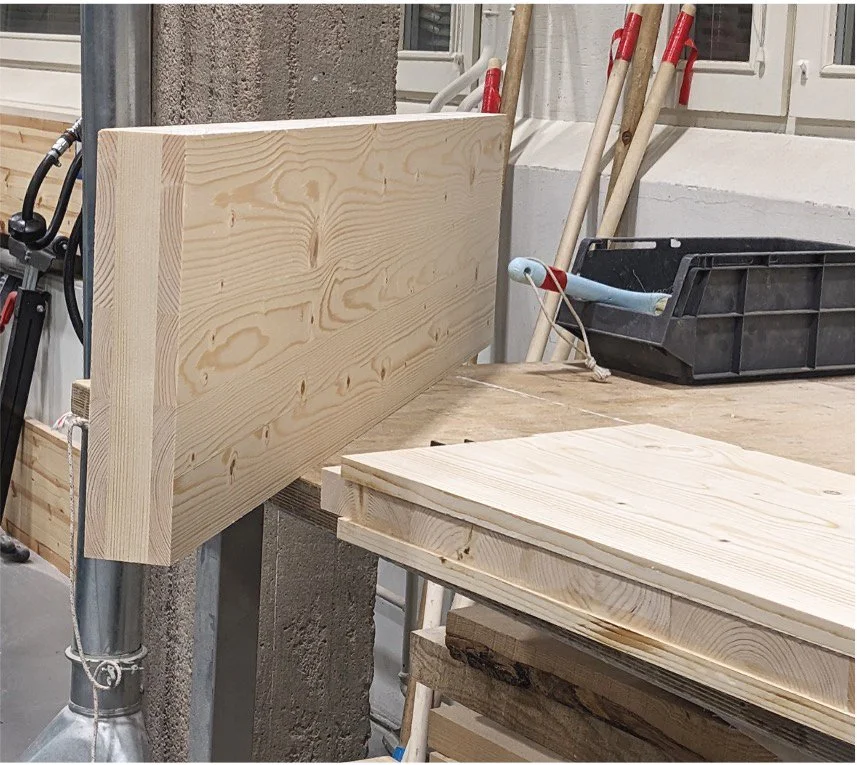 A woodworking workshop with a wooden board attached to a wall, a workbench with additional wooden boards, and tools like a saw and a black storage box in the background.