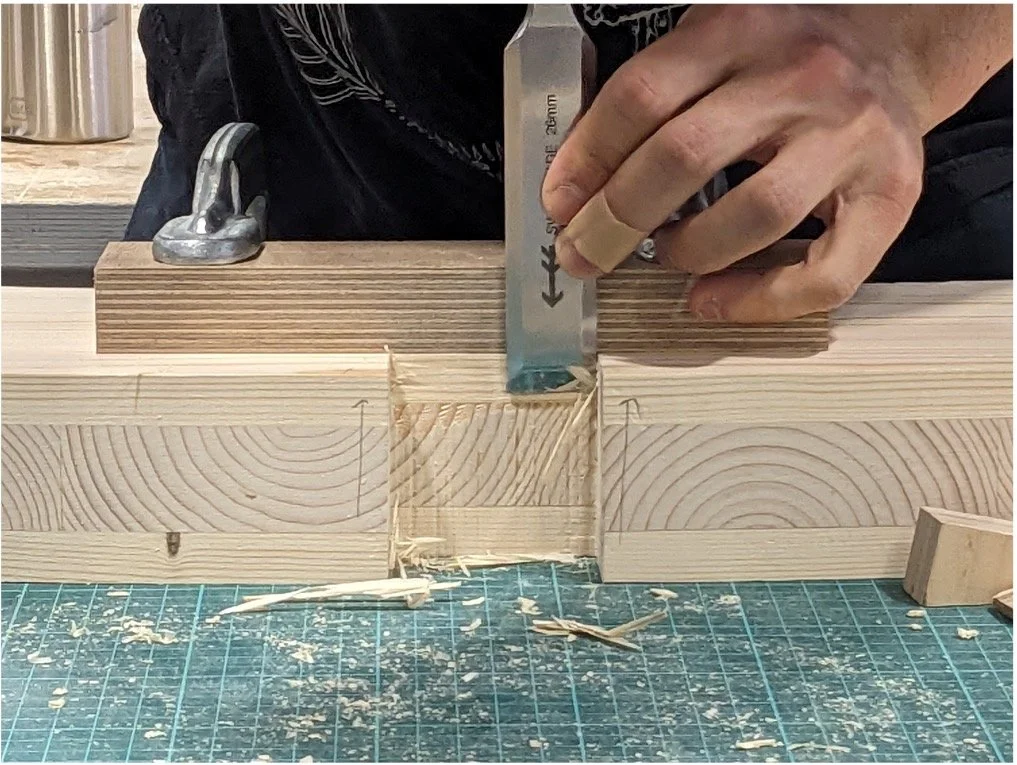 A person is using a chisel to carve a joint in a piece of wood during woodworking. Wood shavings are scattered on a green cutting mat on the workbench.