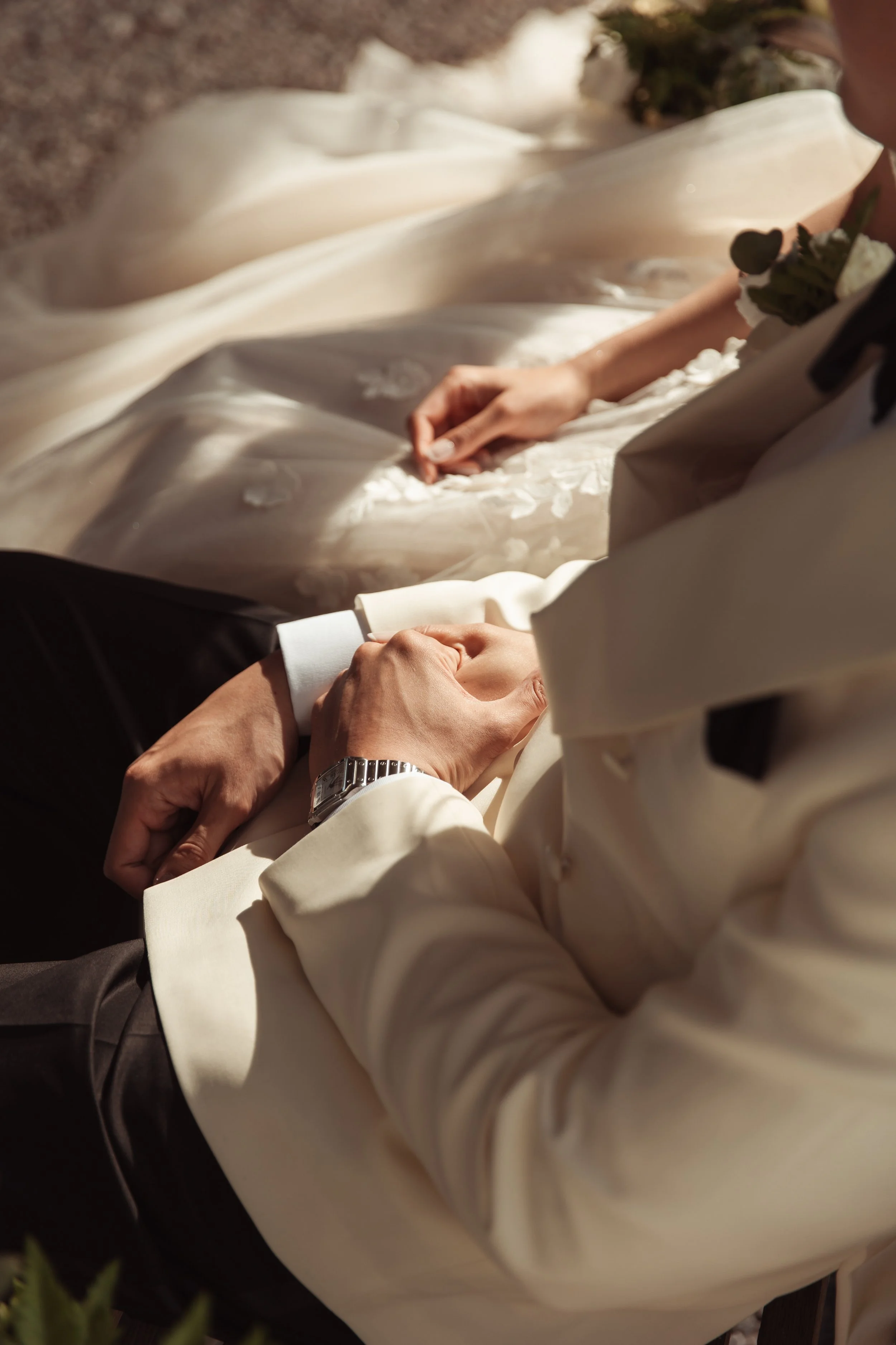 Due persone, probabilmente sposi, si tengono per mano mentre sono seduti, con abiti eleganti, in un ambiente luminoso e raffinato.