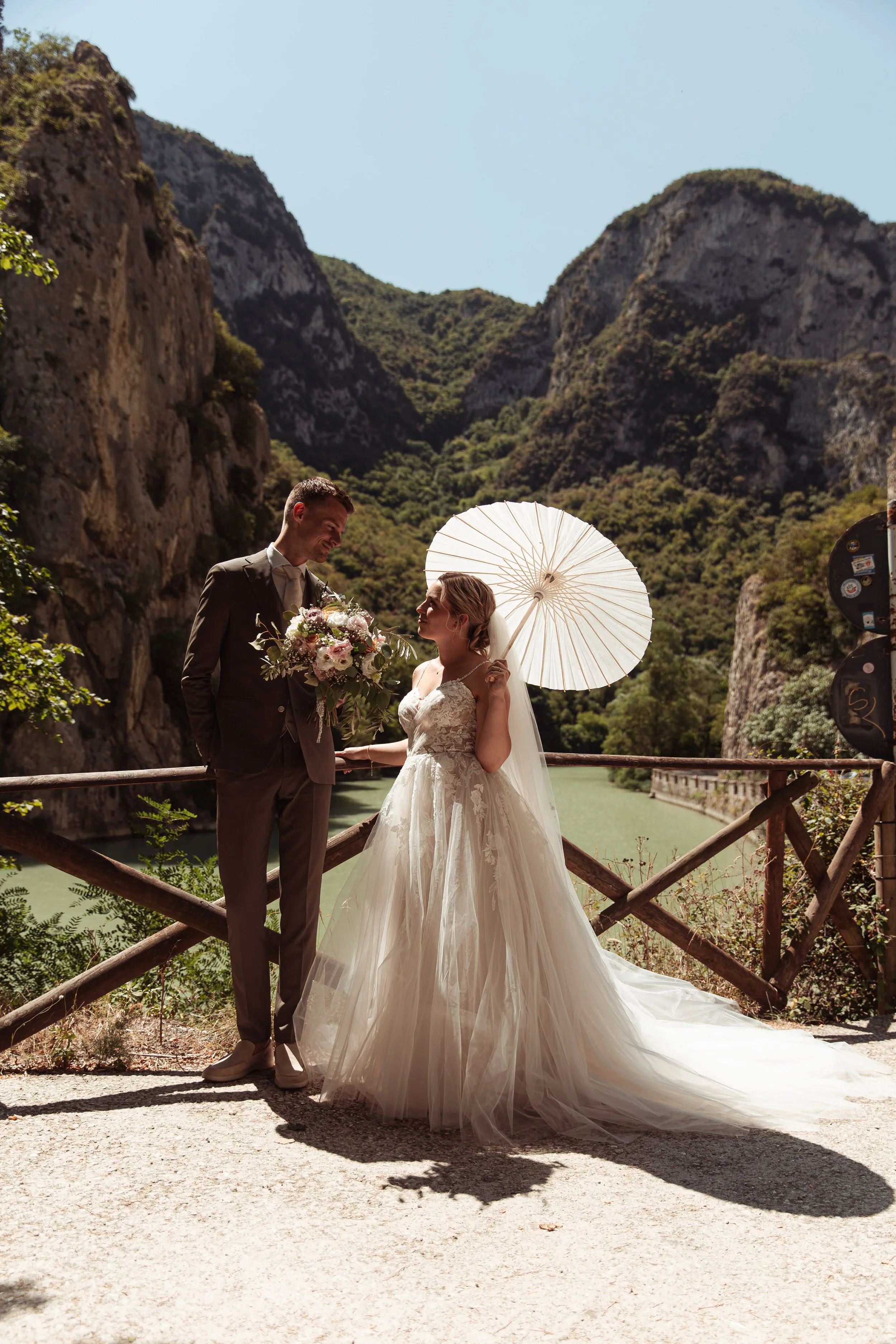 Una coppia di sposi in abiti eleganti si tiene per mano sotto un ombrello bianco in un paesaggio montano con lago e scogliere.