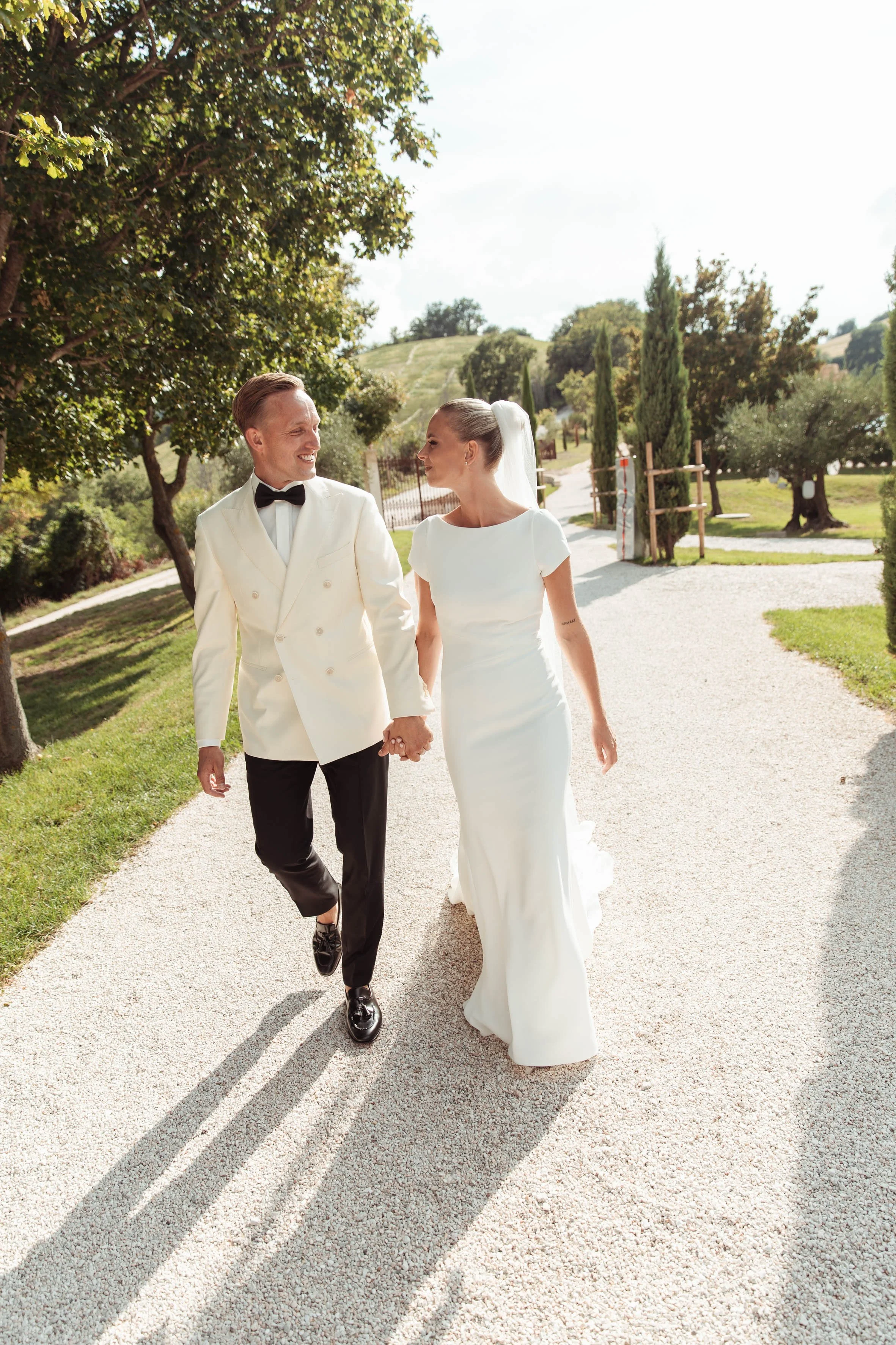 Una coppia di sposi cammina mano nella mano all'aperto, con alberi e natura sullo sfondo in una giornata soleggiata.