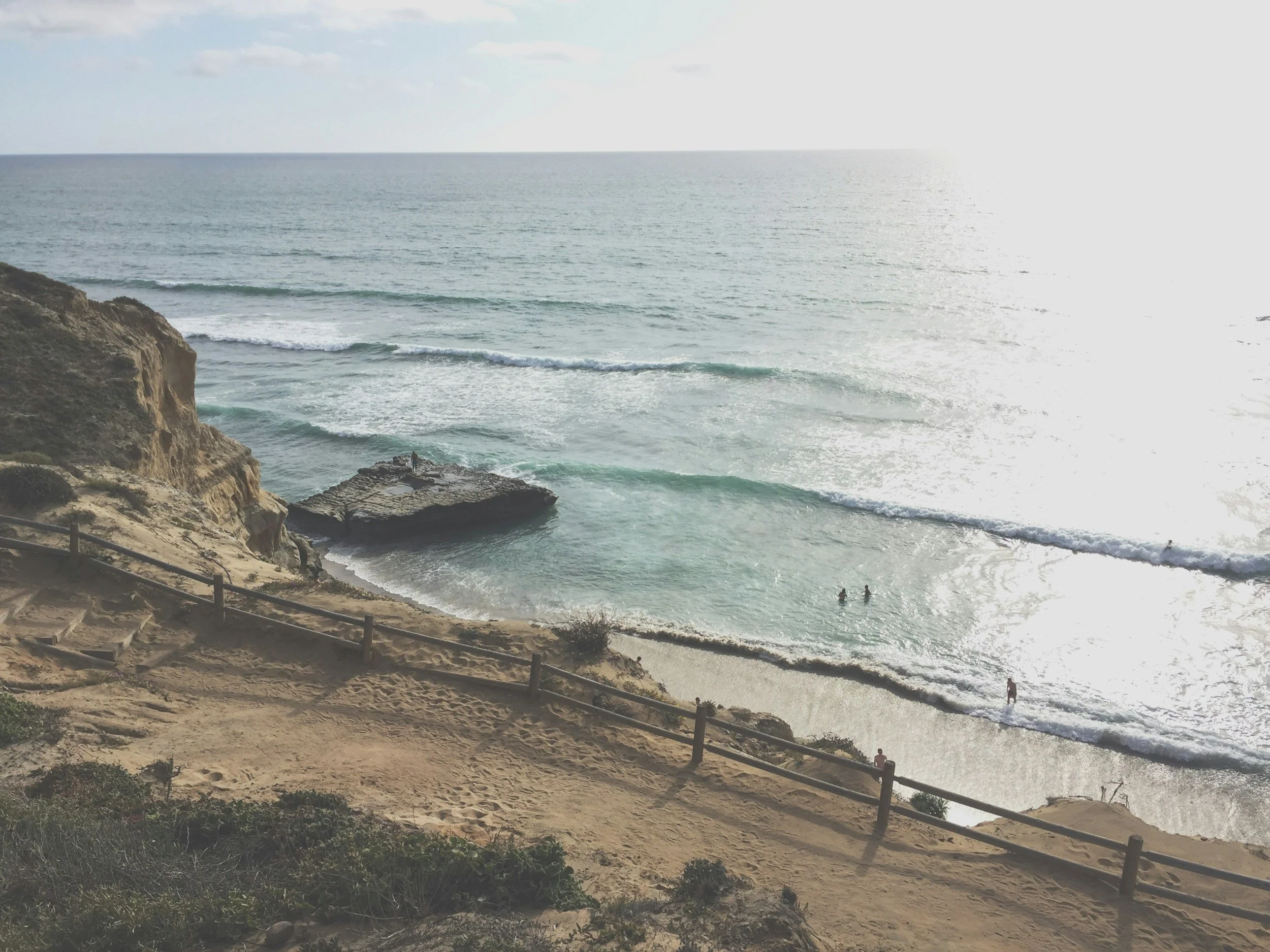 Coastal view of a sandy beach, cliffs, and the ocean with waves, with people swimming and standing near the shore, and a rock formation extending into the water.