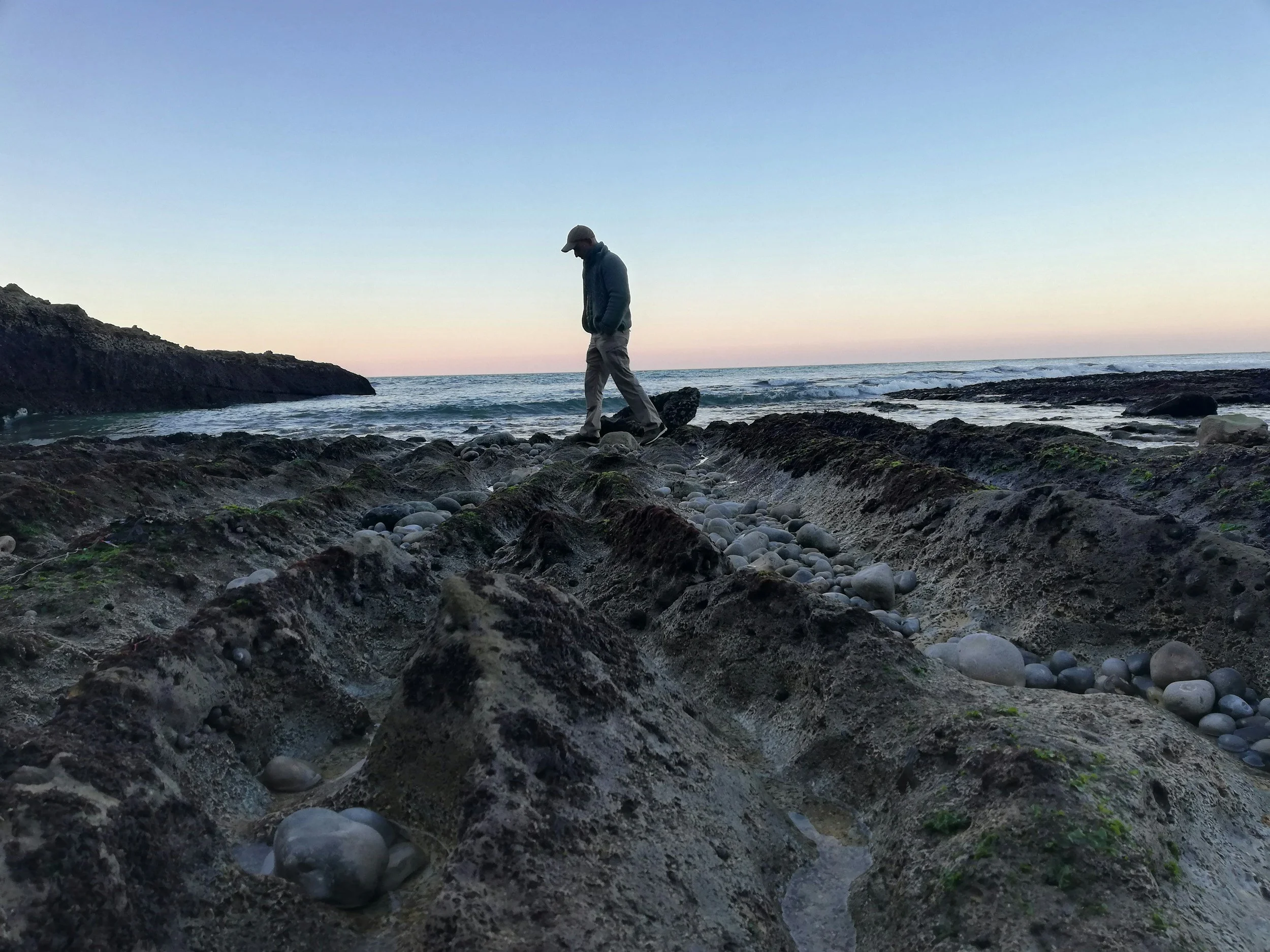 A person walking along a rocky beach during sunset or sunrise, with the ocean and sky in the background.