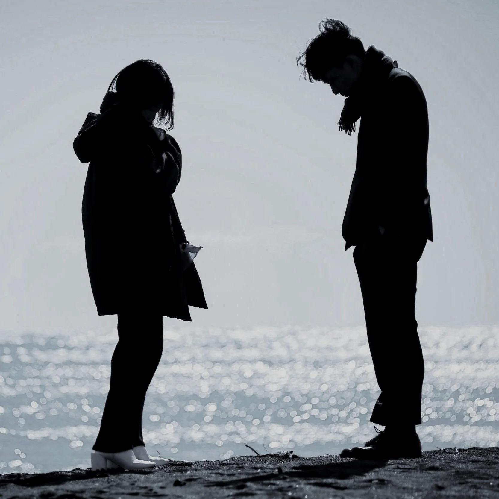 Silhouettes of a man and a woman with relationship issues standing and looking down at the ground near the water on a beach, with the ocean shimmering in the background.