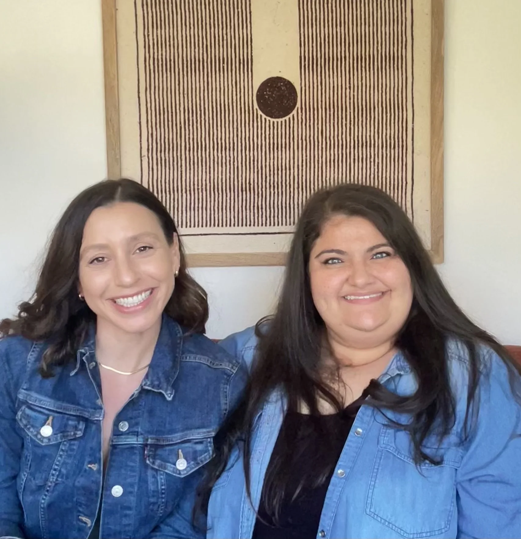 Two women sitting together, smiling at the camera, wearing denim jackets, with abstract wall art behind them.