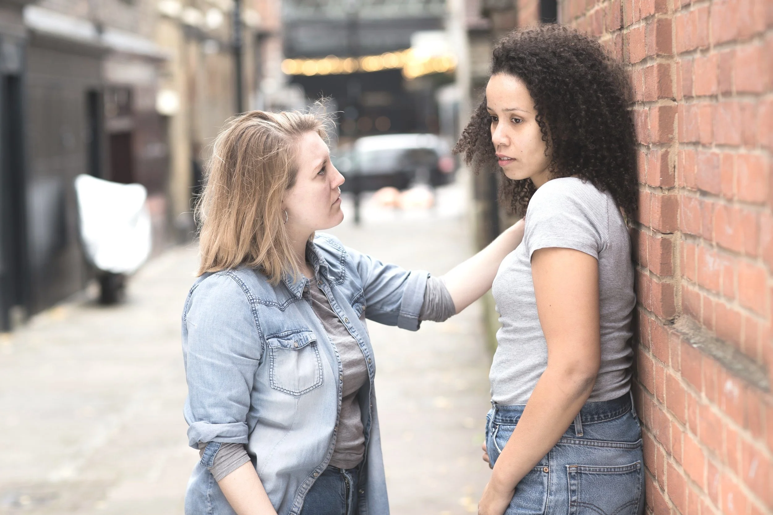 A lesbian couple with relationship issues having an intense conversation on a city street, one woman leaning against a brick wall, the other emotionally engaging.