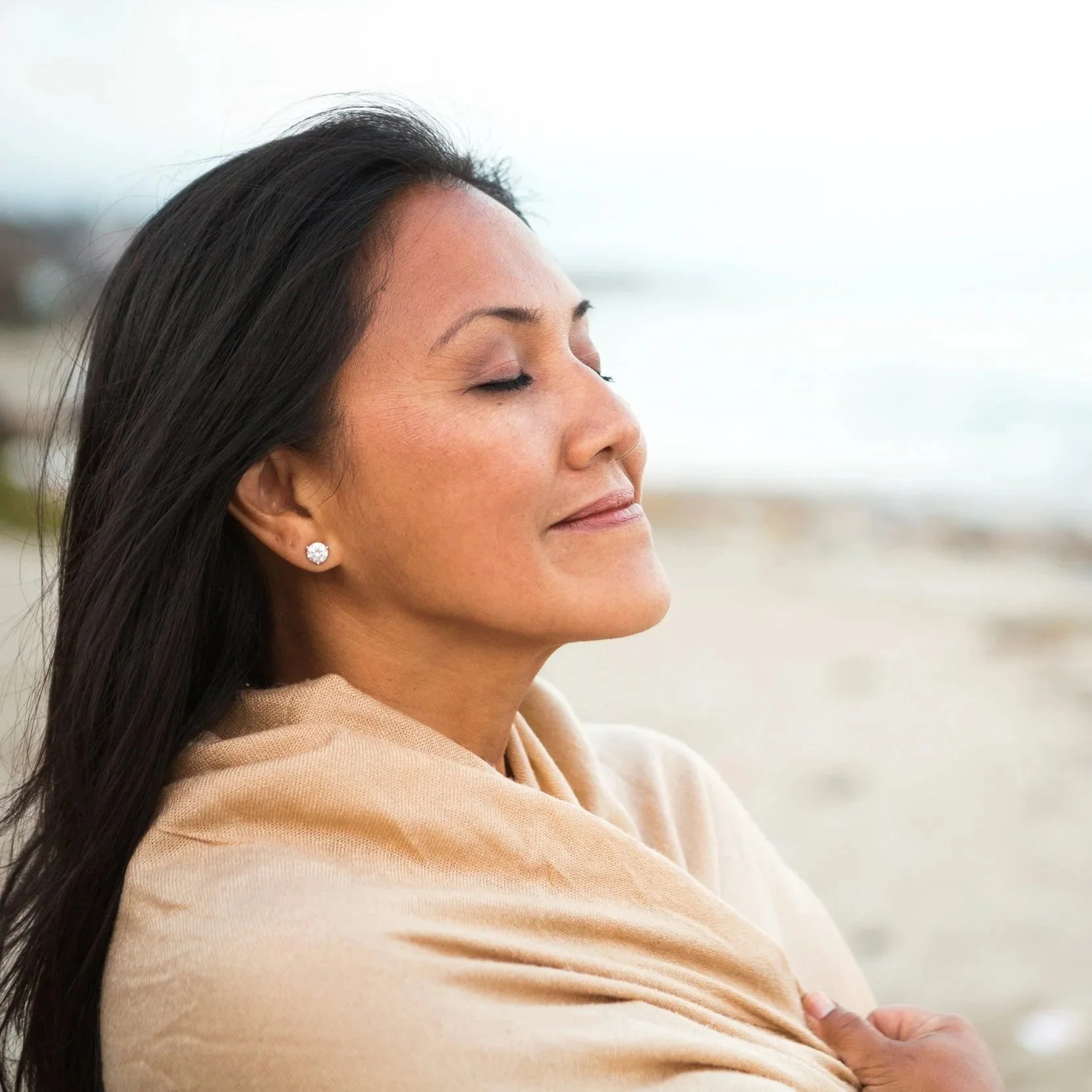 A woman with dark hair and earrings stands outdoors with eyes closed, appearing to enjoy the moment near the beach.