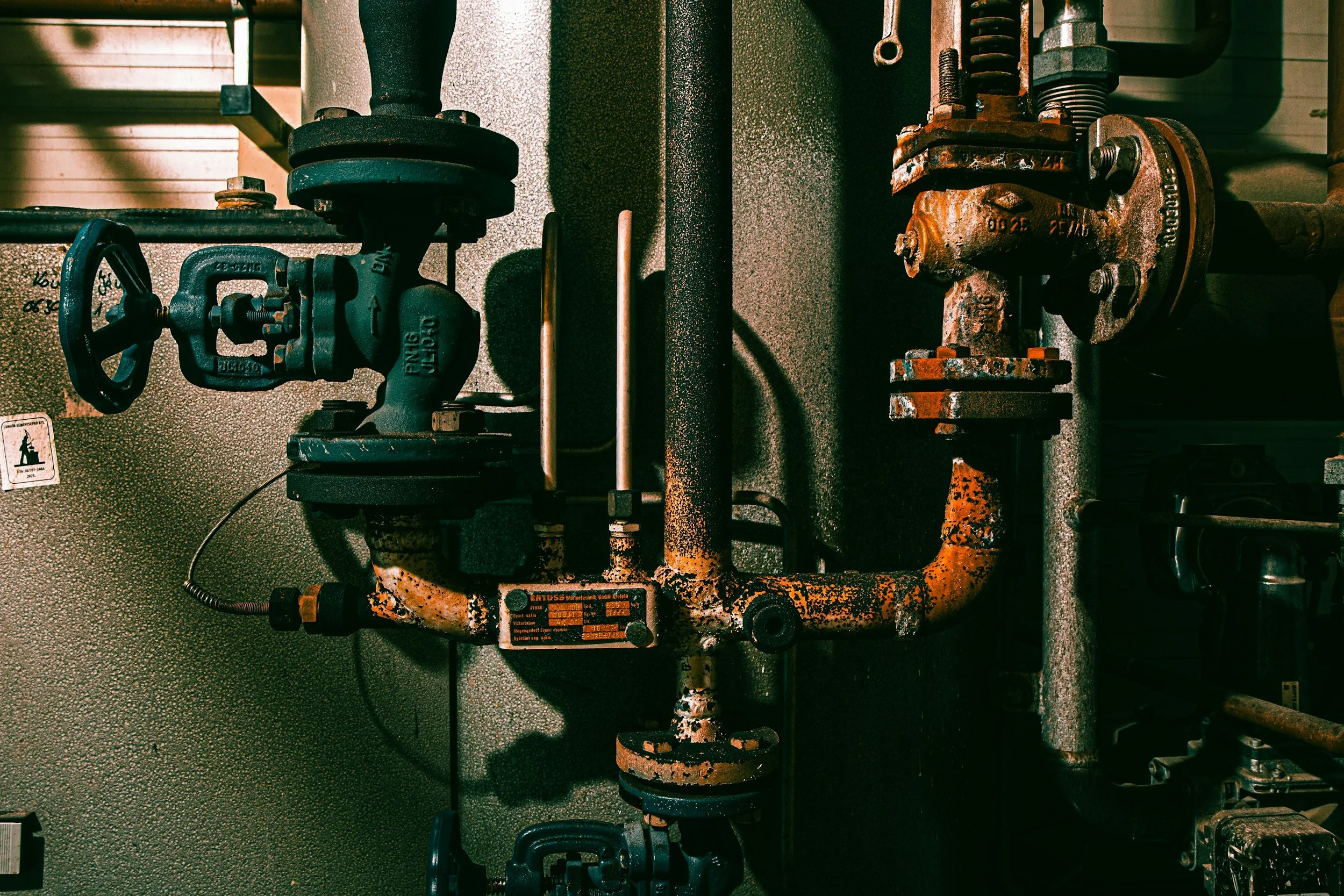 A close-up of an industrial piping system with a rusted orange pipe, a black valve, and other metal components on a textured grey surface.