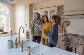Three people standing in a kitchen, looking at a tablet together, with a vintage stove and window in the background.