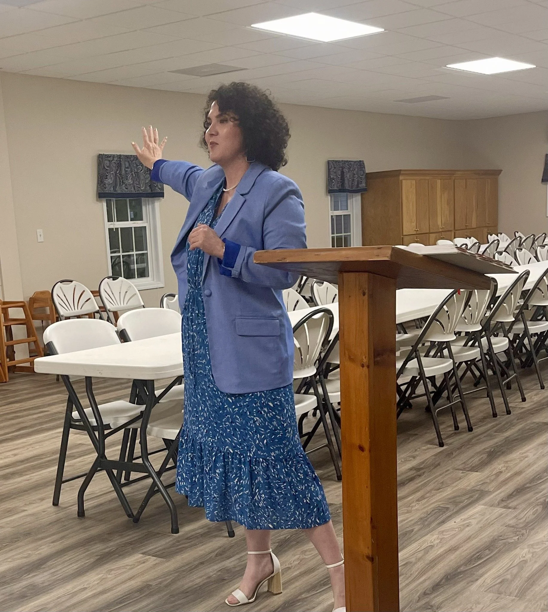 Woman speaking at a podium in a room with white tables and chairs, wearing a blue dress and blazer, with curly hair and heels.