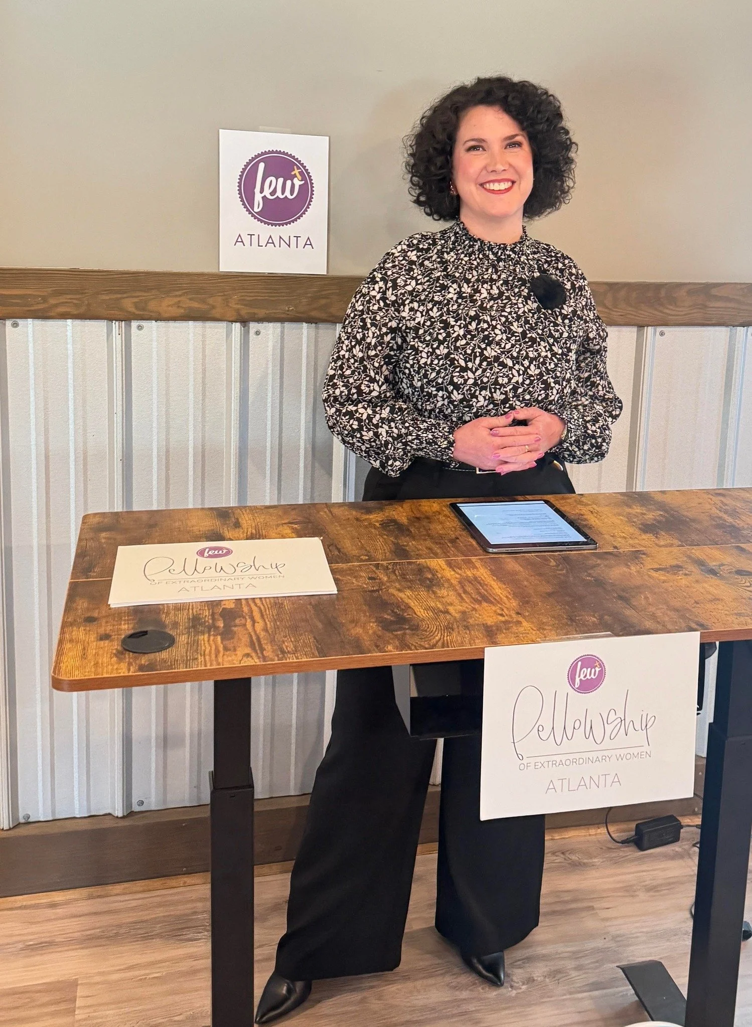 A woman with curly hair standing behind a wooden table with a sign that reads 'Fellowship of Extraordinary Women ATLANTA.' She is smiling, wearing a patterned blouse and black pants. On the table, there is a tablet and a paper with a similar sign. In the background, a wall has a purple and white sign that says 'feW ATLANTA.'