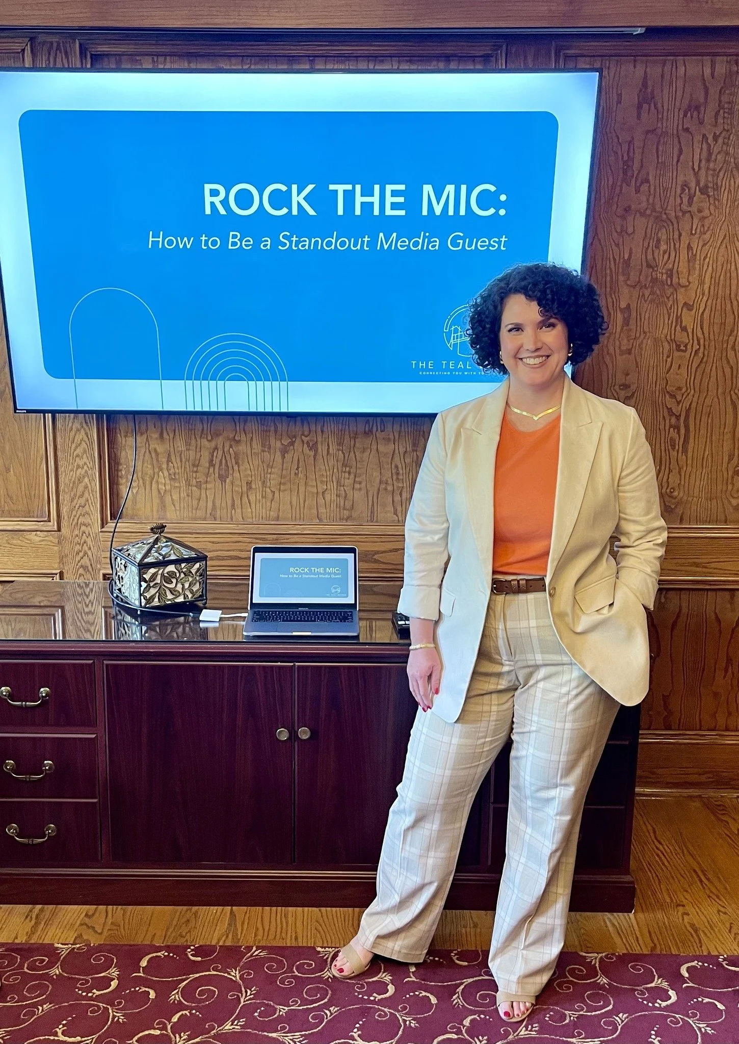 A woman with curly hair wearing a beige blazer, plaid beige pants, and a bright orange top, standing in front of a large screen and smiling. The screen displays a presentation titled 'Rock the Mic: How to Be a Standout Media Guest.' There is a laptop on a wooden cabinet and a small decorative lamp beside it.