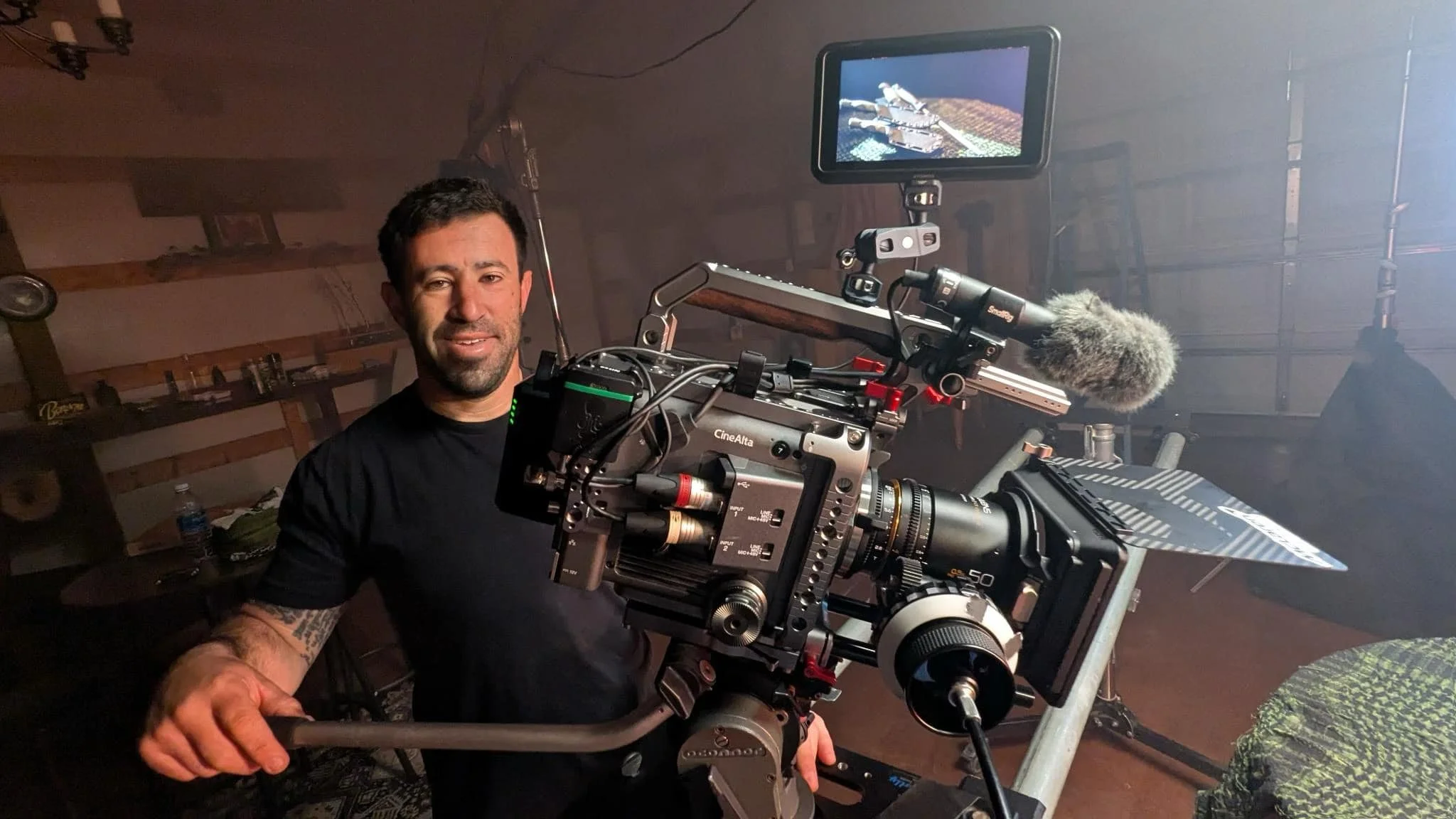 Man with dark hair and beard smiling behind professional film camera in a dimly lit room.