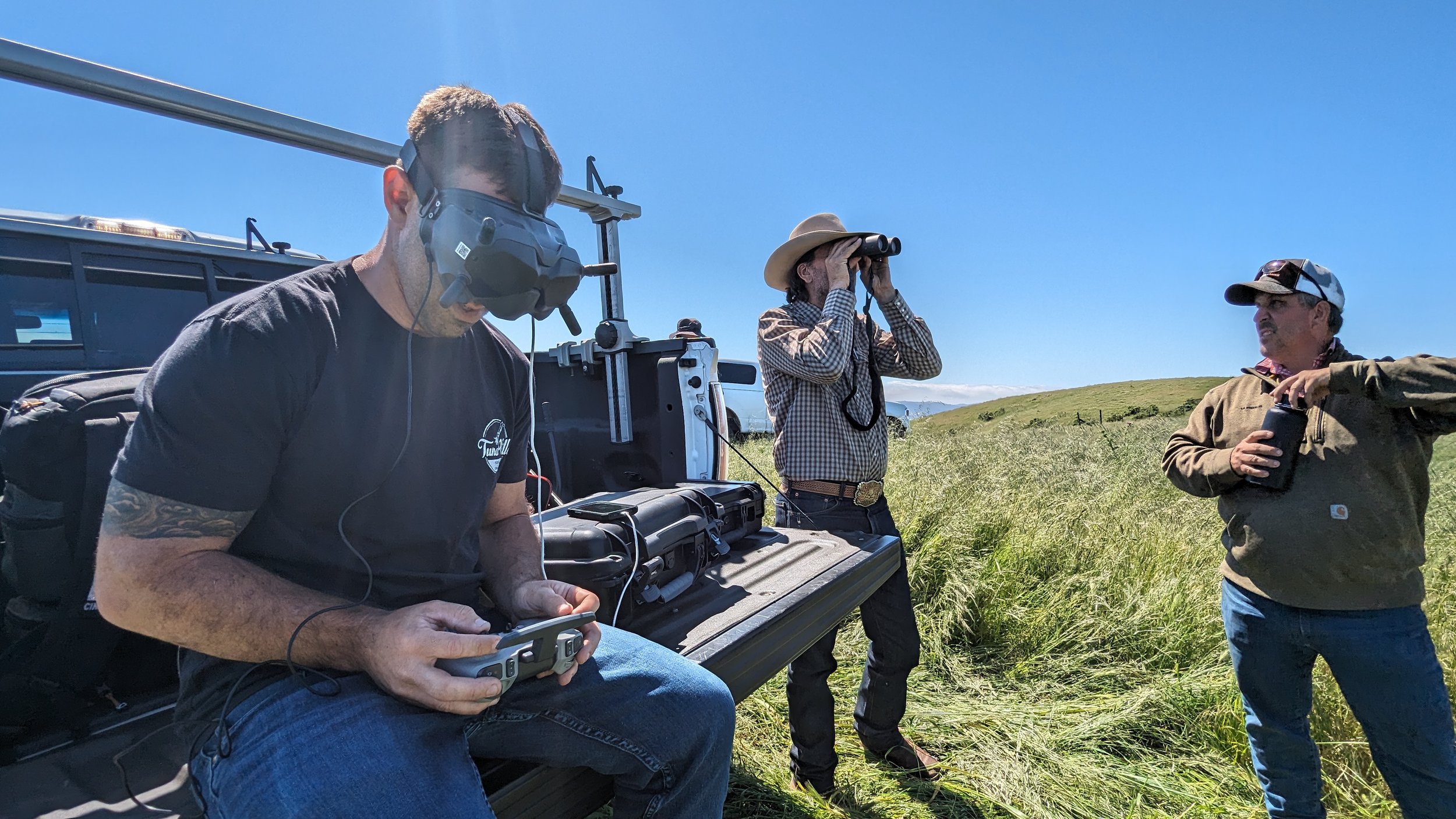 Three men in a grassy field with clear blue sky, some vehicles in the background. One man is wearing a drone headset and holding a controller, another is looking through binoculars, and the third is holding a drink and gesturing.