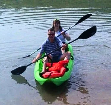 Two people are paddling a green kayak on calm water, surrounded by a serene natural landscape. Life jackets are visible in the kayak.