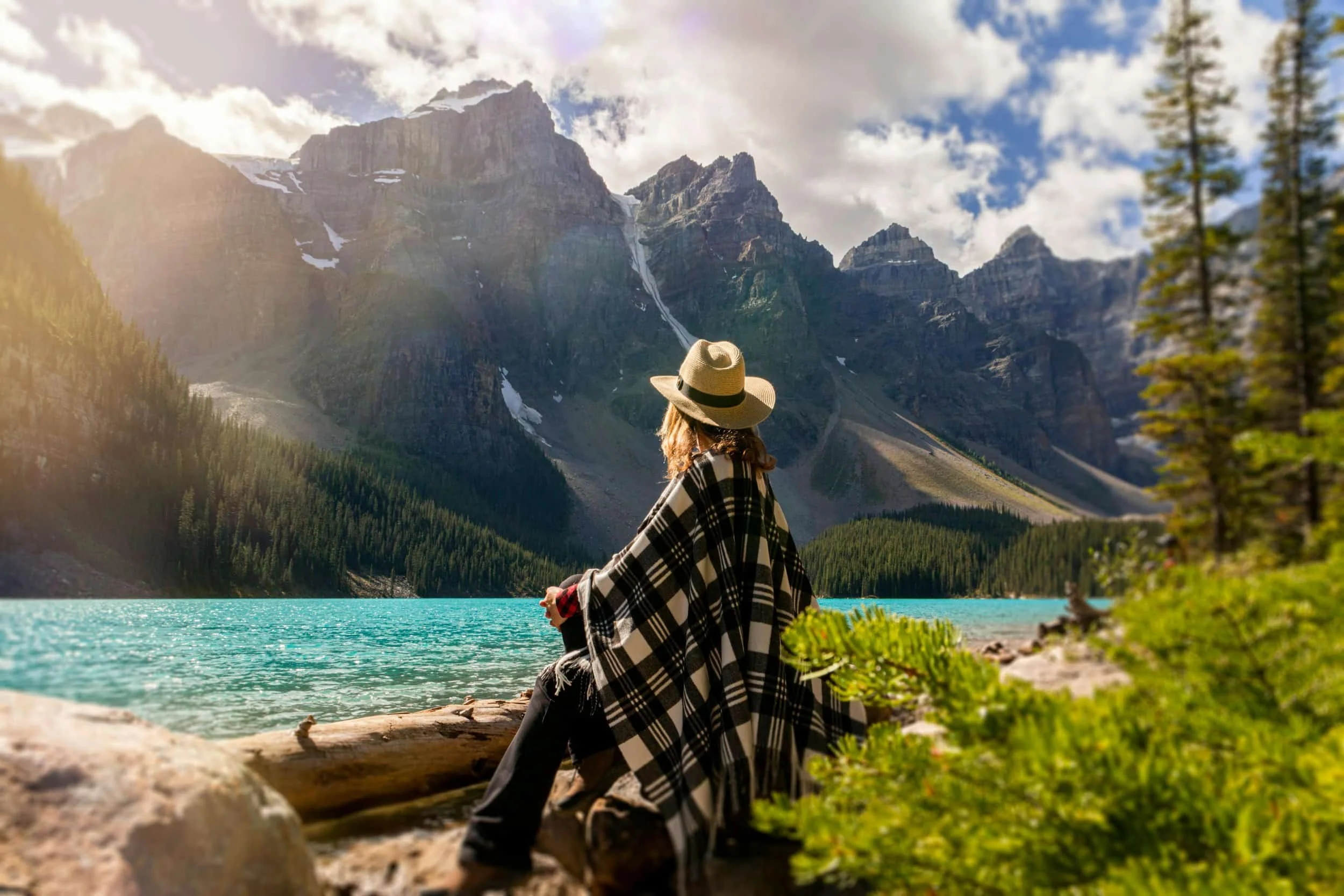 A woman wearing a hat and a checkered poncho sits on the side of a beautiful mountain lake at peace