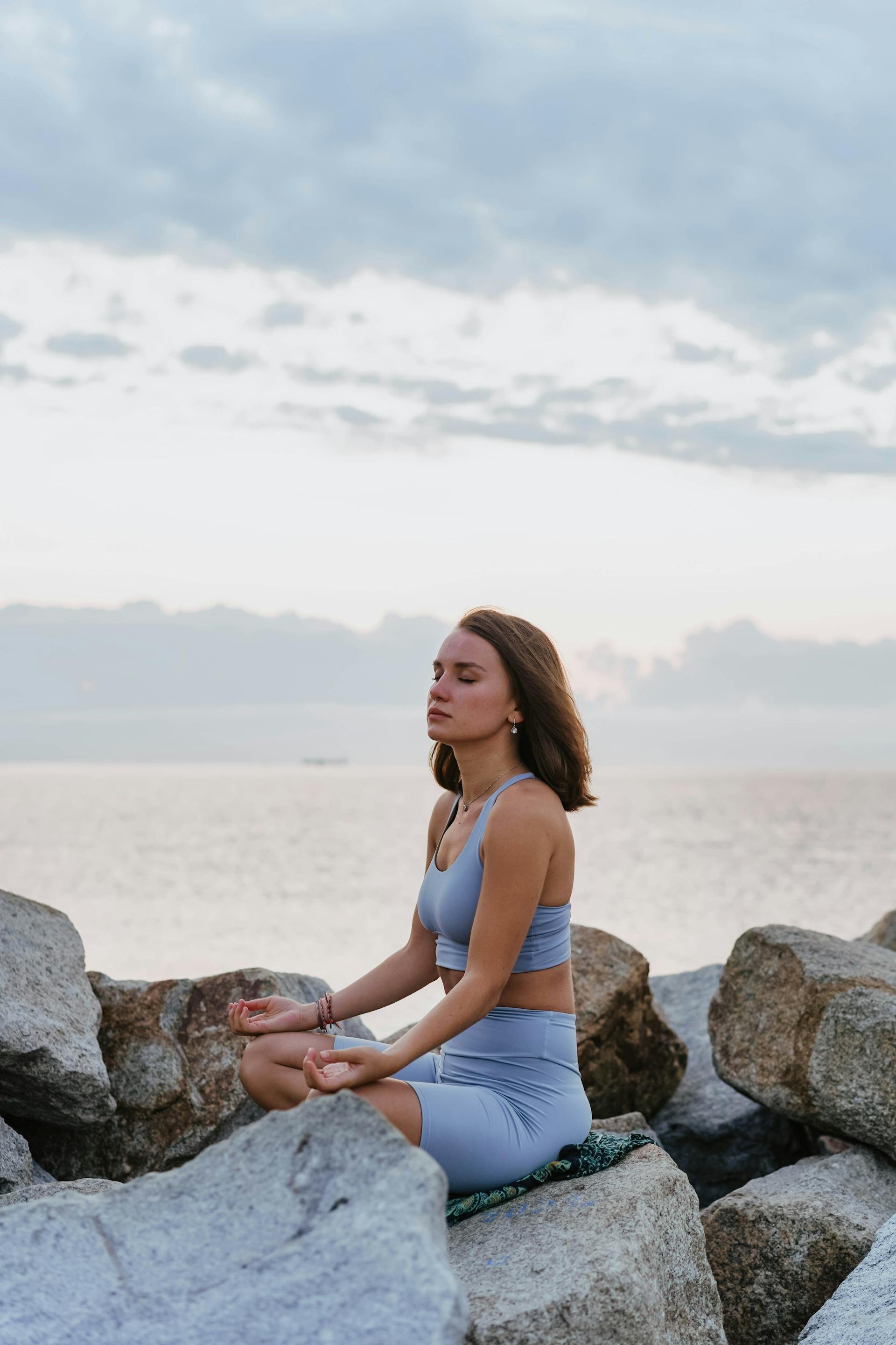 A woman practicing yoga outdoors on large rocks near a body of water, with her eyes closed and her hands resting on her knees in a meditative pose.
