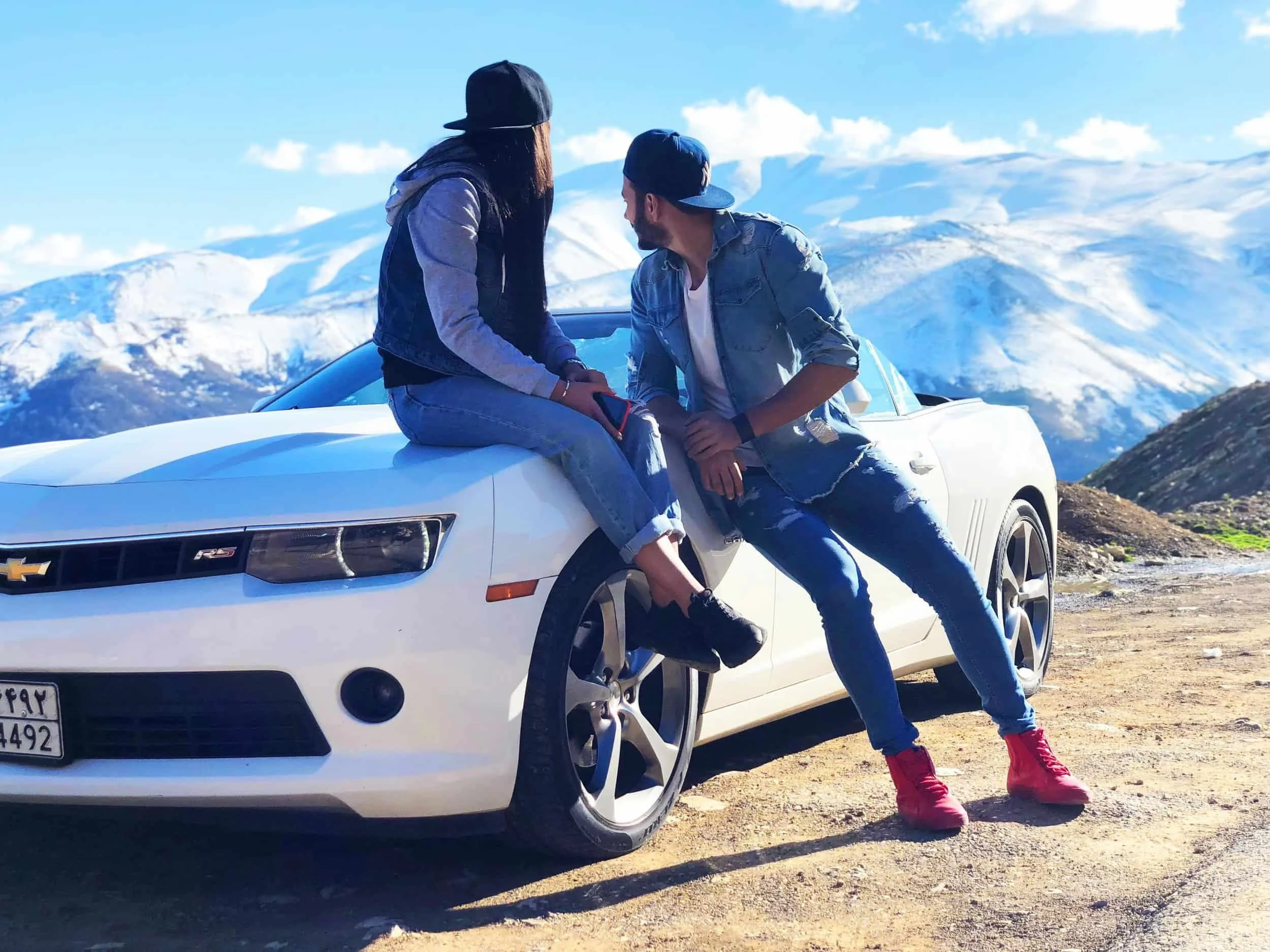 Two people in casual clothes sit and lean on a white sports car with snowy mountains in the background under a clear sky.