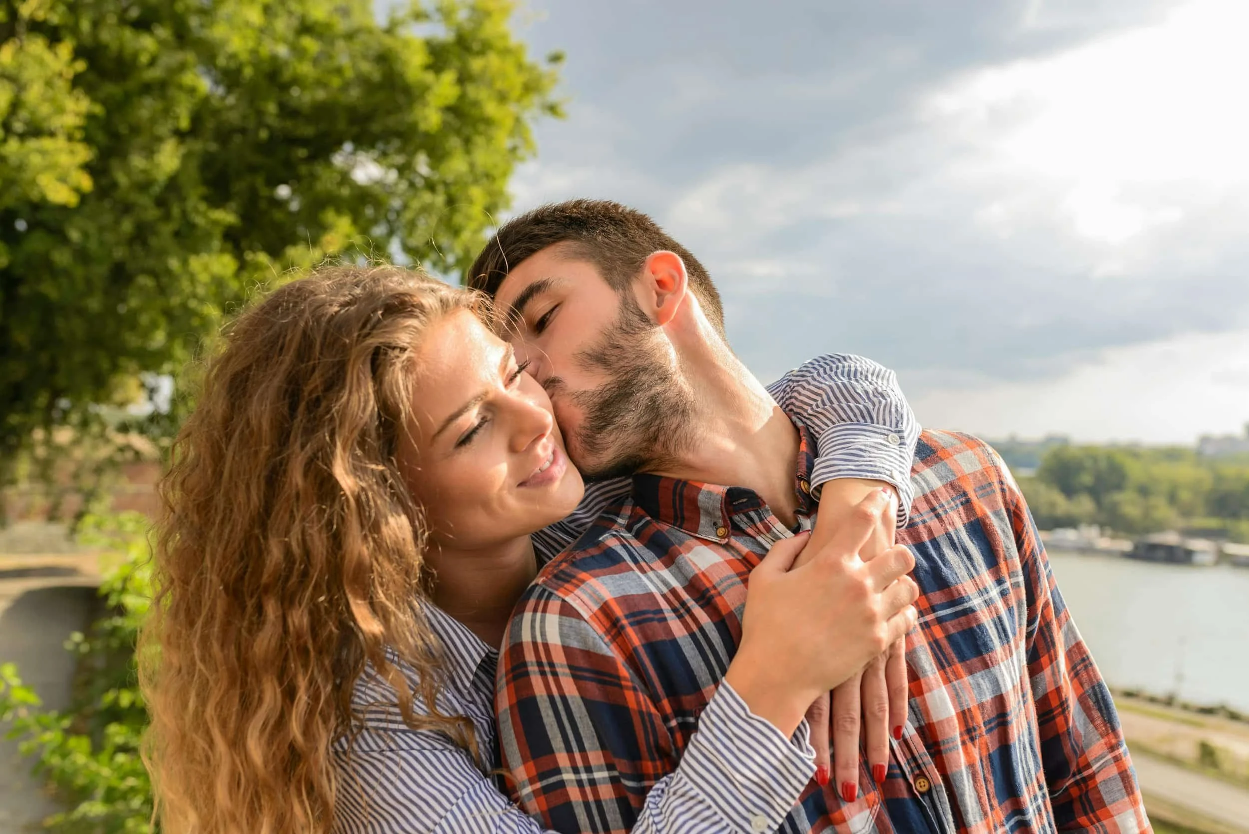 Couple embracing outdoors by a river, with trees and a cloudy sky in the background.