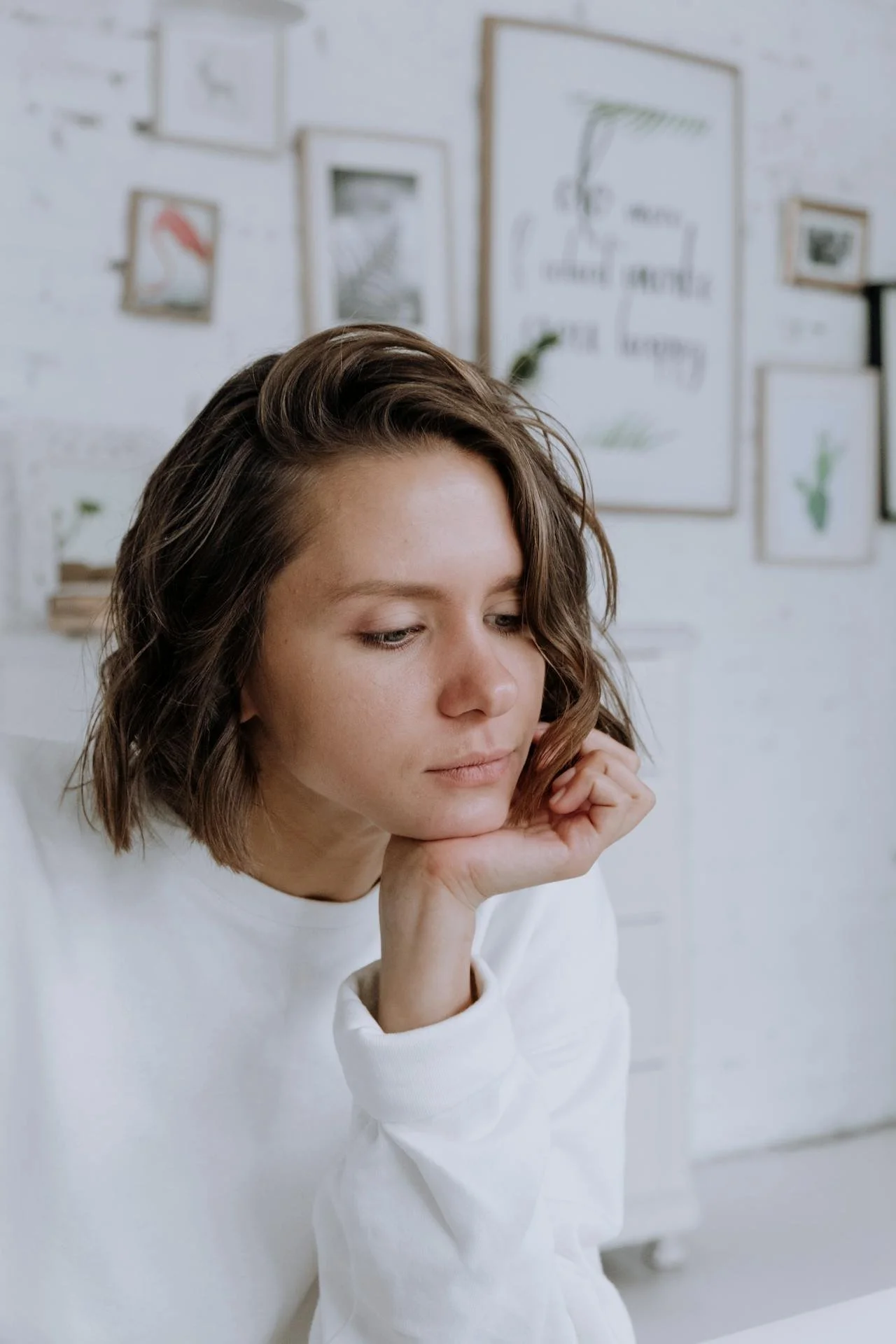 A young woman with shoulder-length brown hair looking down thoughtfully, resting her chin on her hand in front of a white wall decorated with framed artwork.