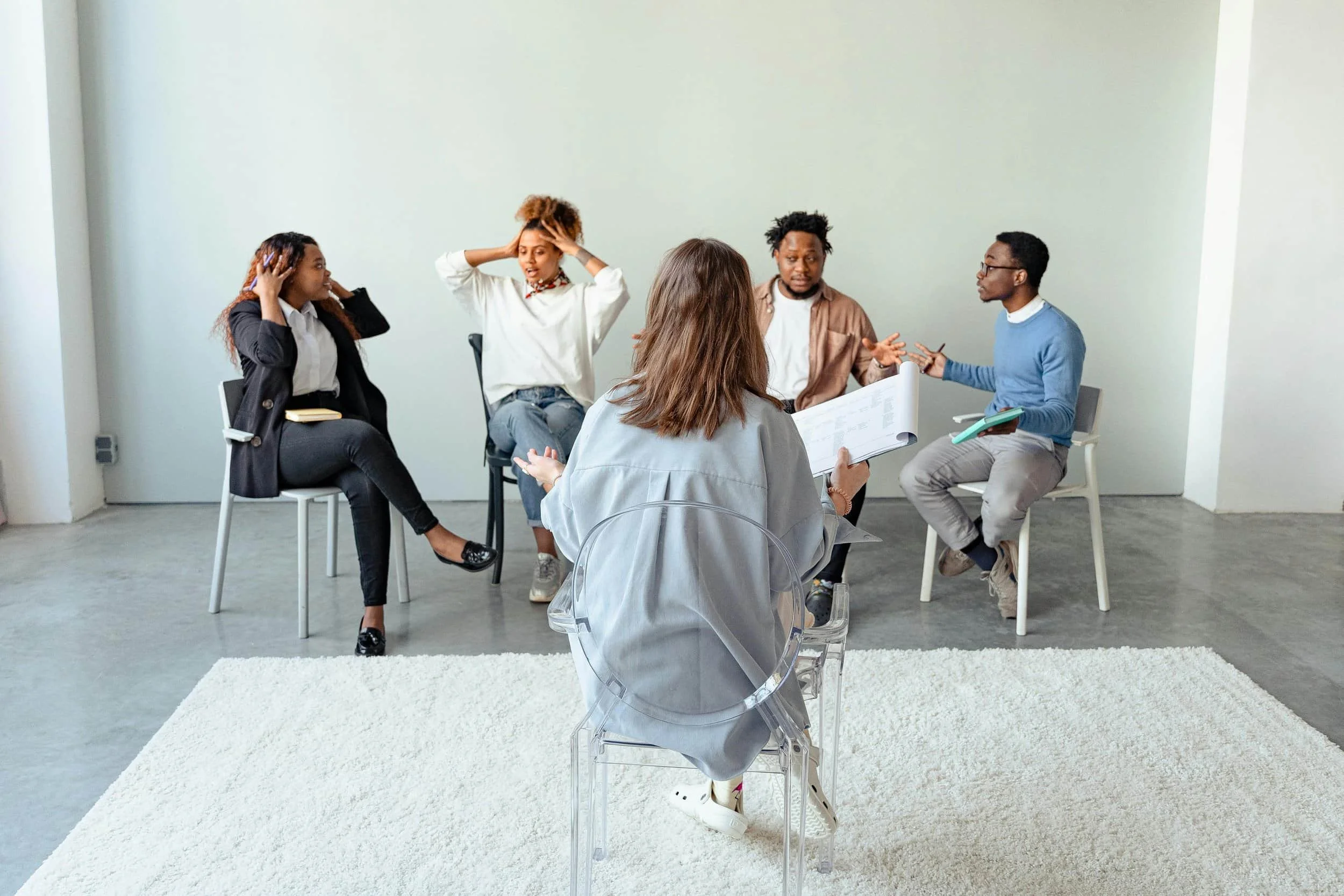 A group discussion in a minimalist space, with participants seated in chairs, engaging in conversation while facing a facilitator.