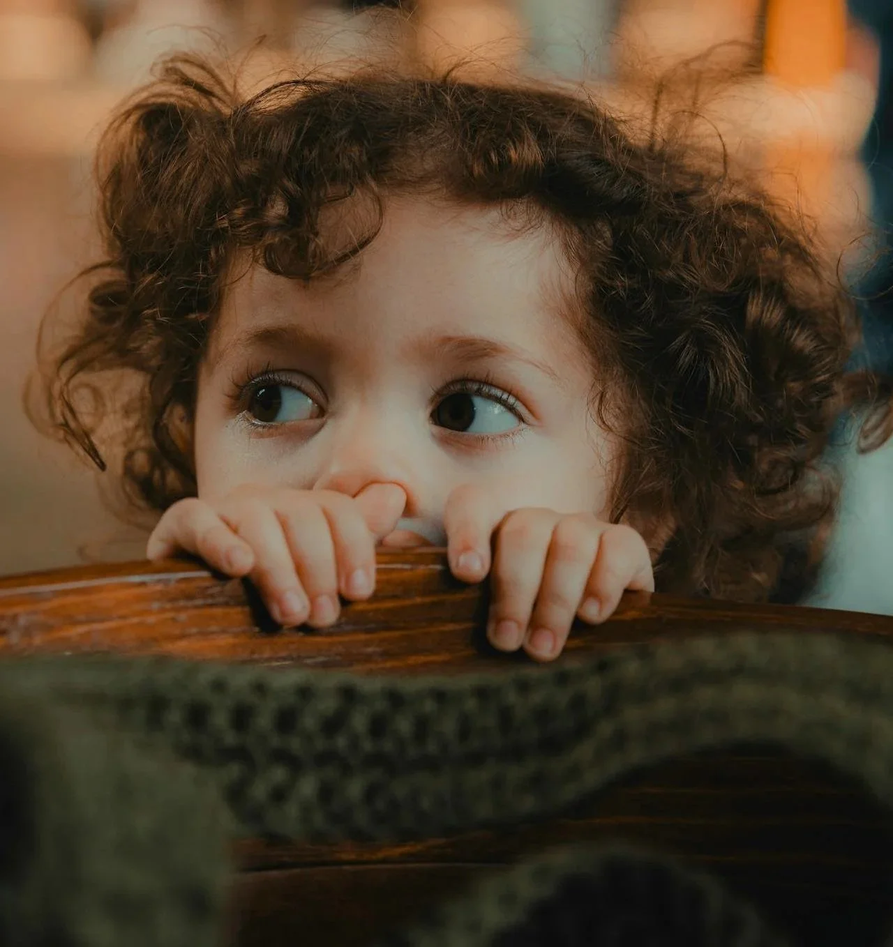 Child with curly hair peeking over the back of a wooden chair against a blurred background with warm lighting.