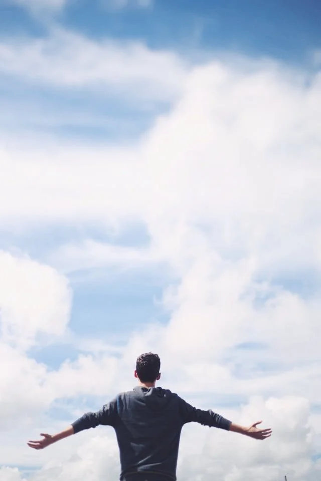 Person standing with arms outstretched under a blue sky filled with fluffy white clouds.