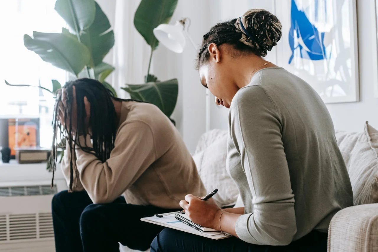 A man sits on a couch in distress holding his head in his hands and therapist sits next to him taking notes