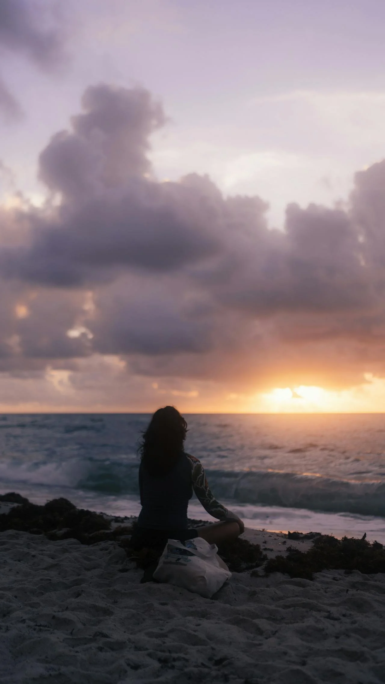 A person sitting on the beach at sunset, facing the ocean with clouds in the sky and the sun setting on the horizon.