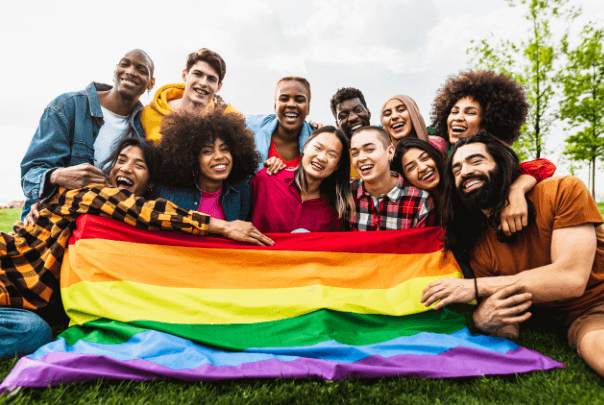 A diverse group of smiling people hold a rainbow pride flag together outdoors on a grassy field.