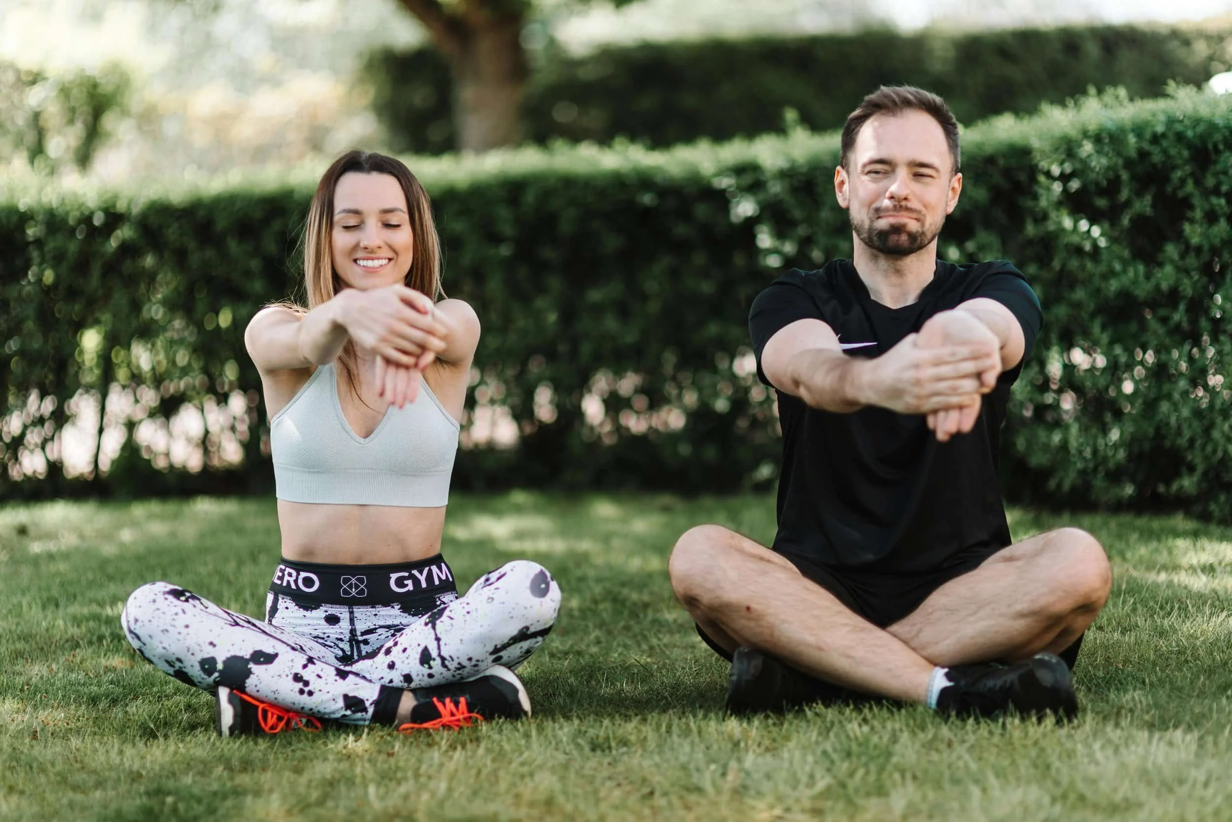 Two people sitting cross-legged on grass, performing an arm stretch outdoors with greenery in the background.