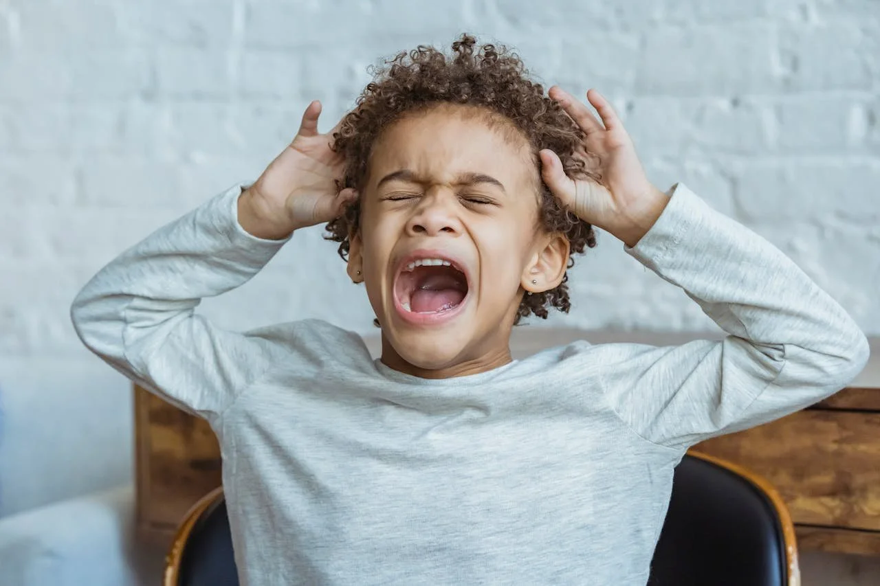 A young child with curly hair is shouting with his eyes closed, holding his head with both hands, in front of a white brick wall.