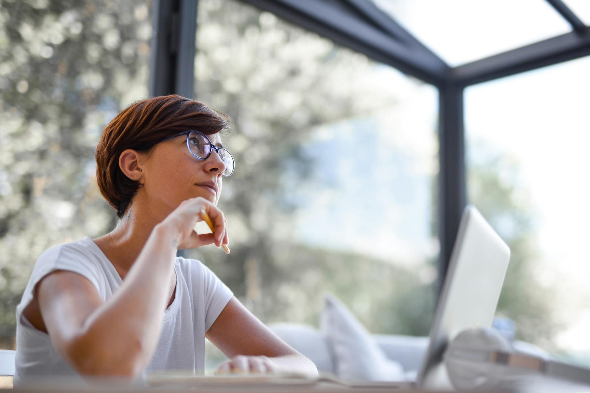 A woman with short brown hair and glasses sitting at a desk, looks contemplative while working on a laptop outdoors near a window with trees in the background.