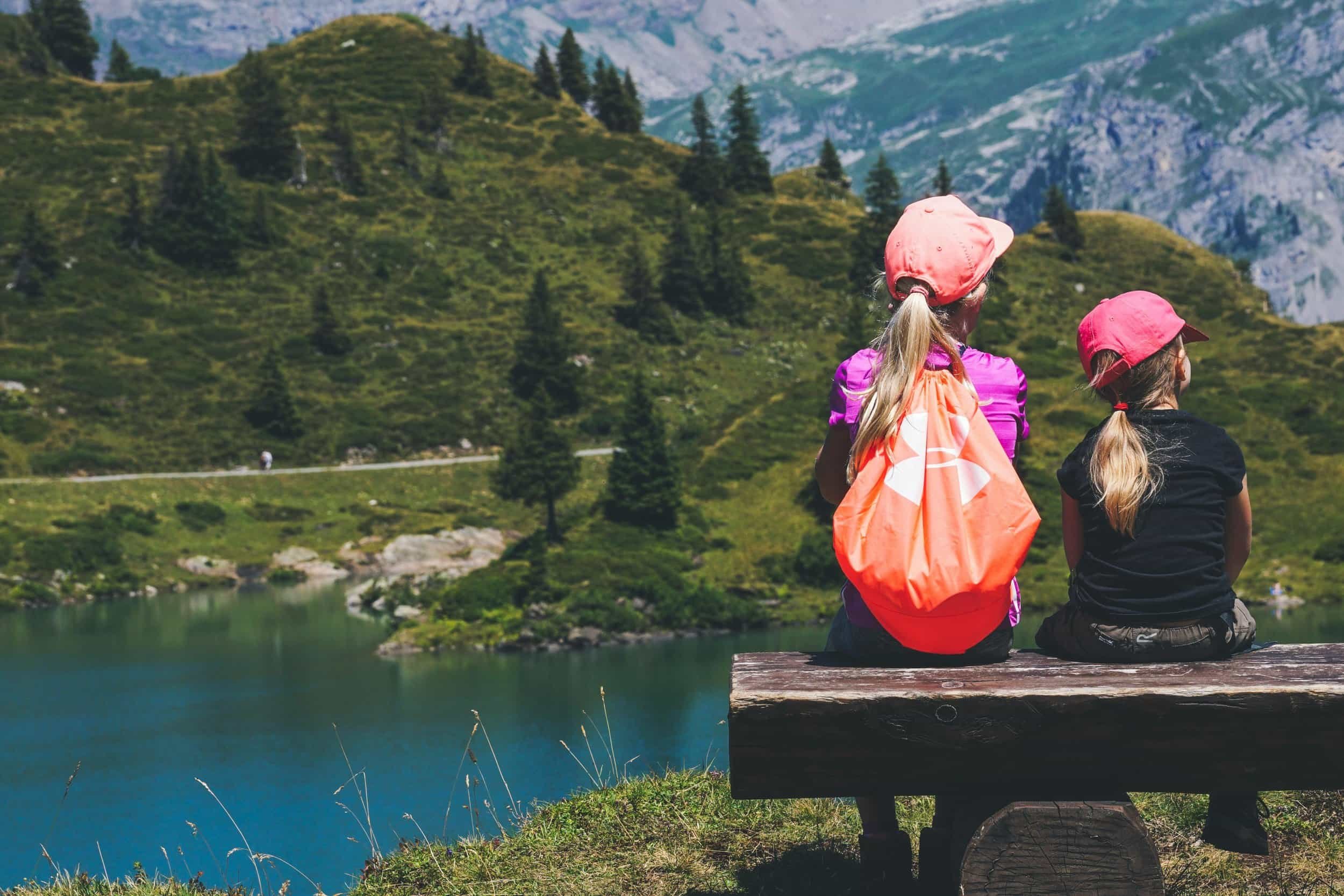 Two children wearing pink baseball caps sit on a bench overlooking a beautiful mountain lake