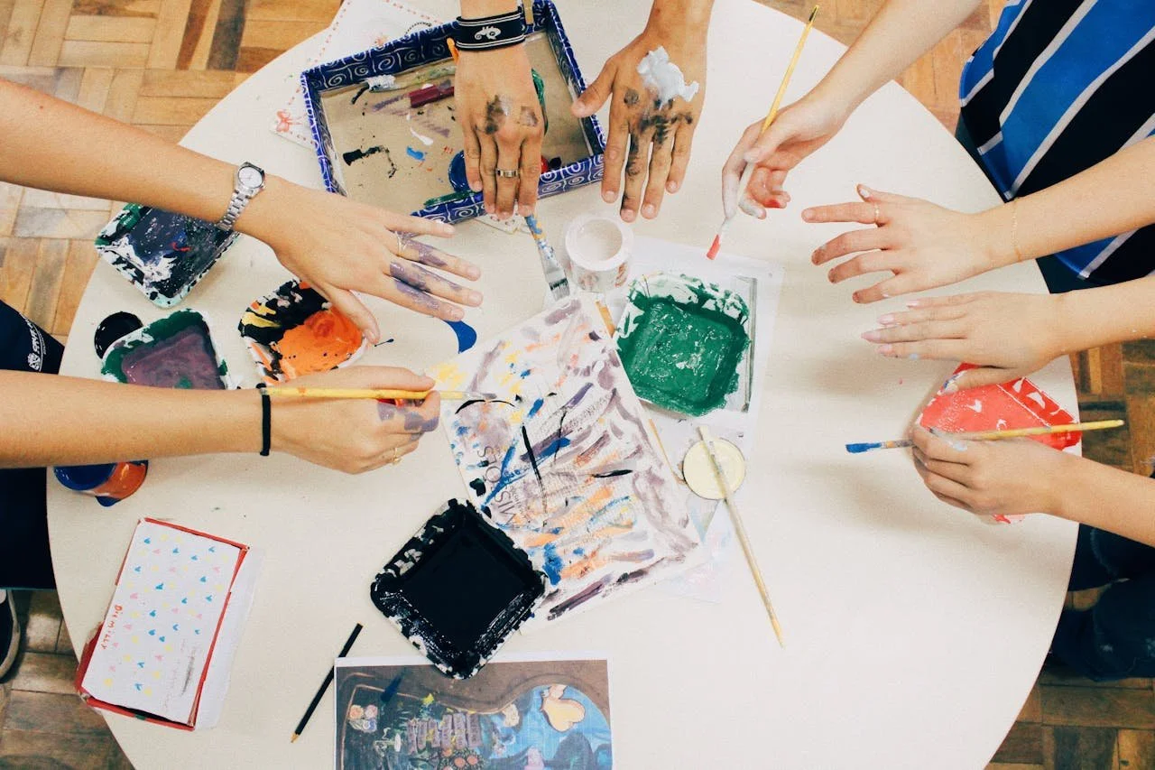 Hands of people painting with brushes and colors on a paper placed on a round table during an art activity.
